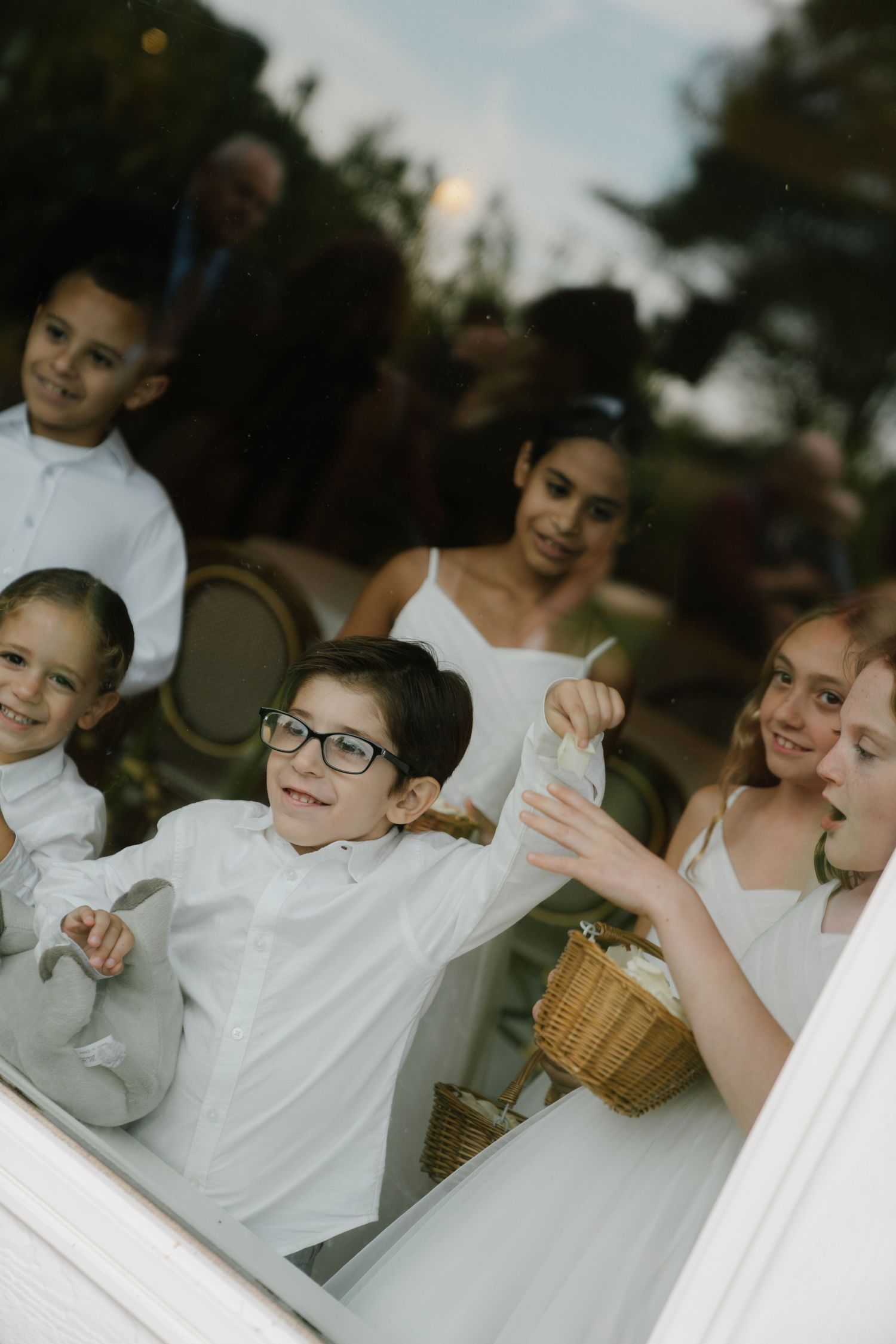 A group of children in white shirts sit together at a gathering, sharing animated expressions and gestures.