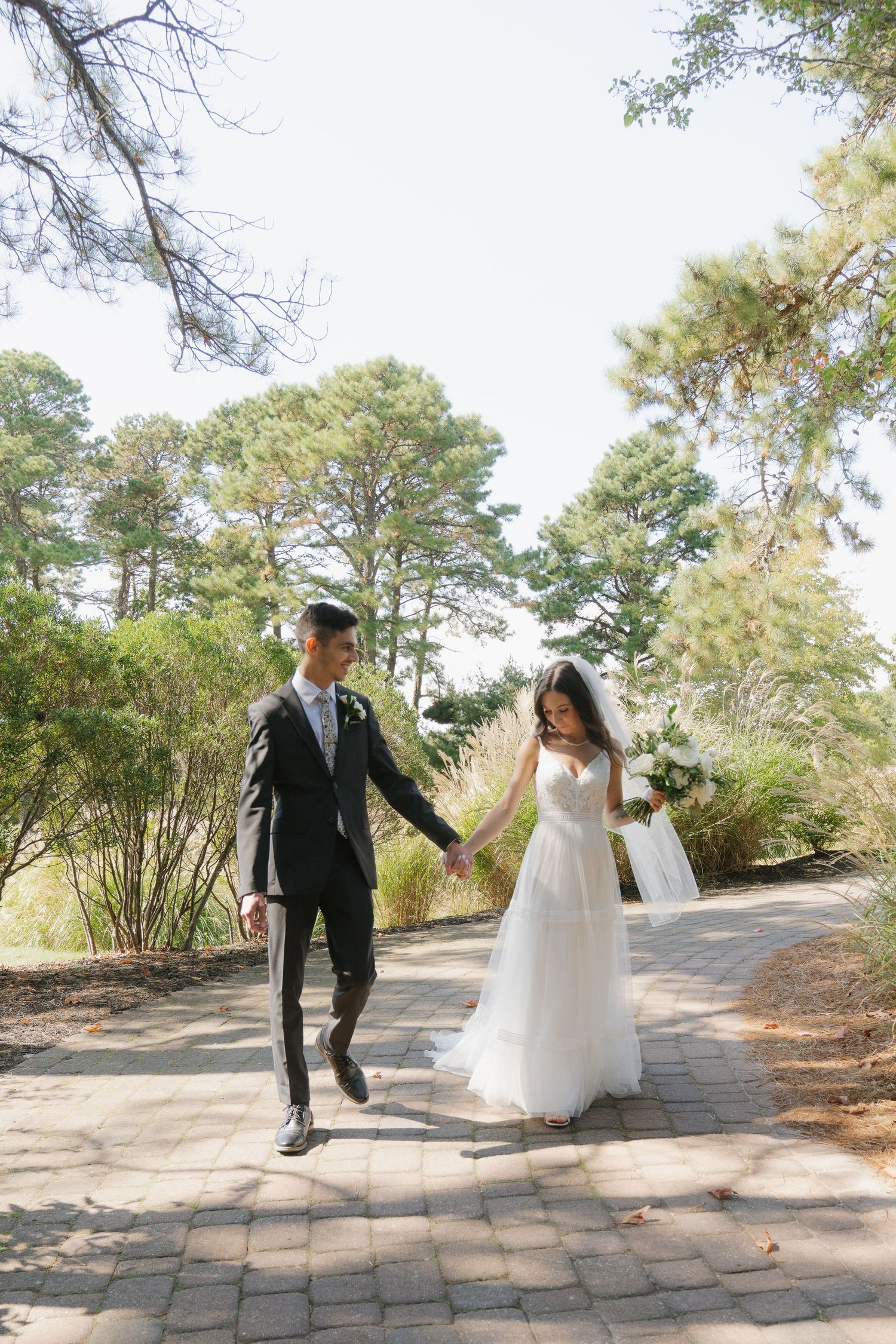 A bride and groom playfully run and dance together on a stone path surrounded by pine trees on their wedding day.