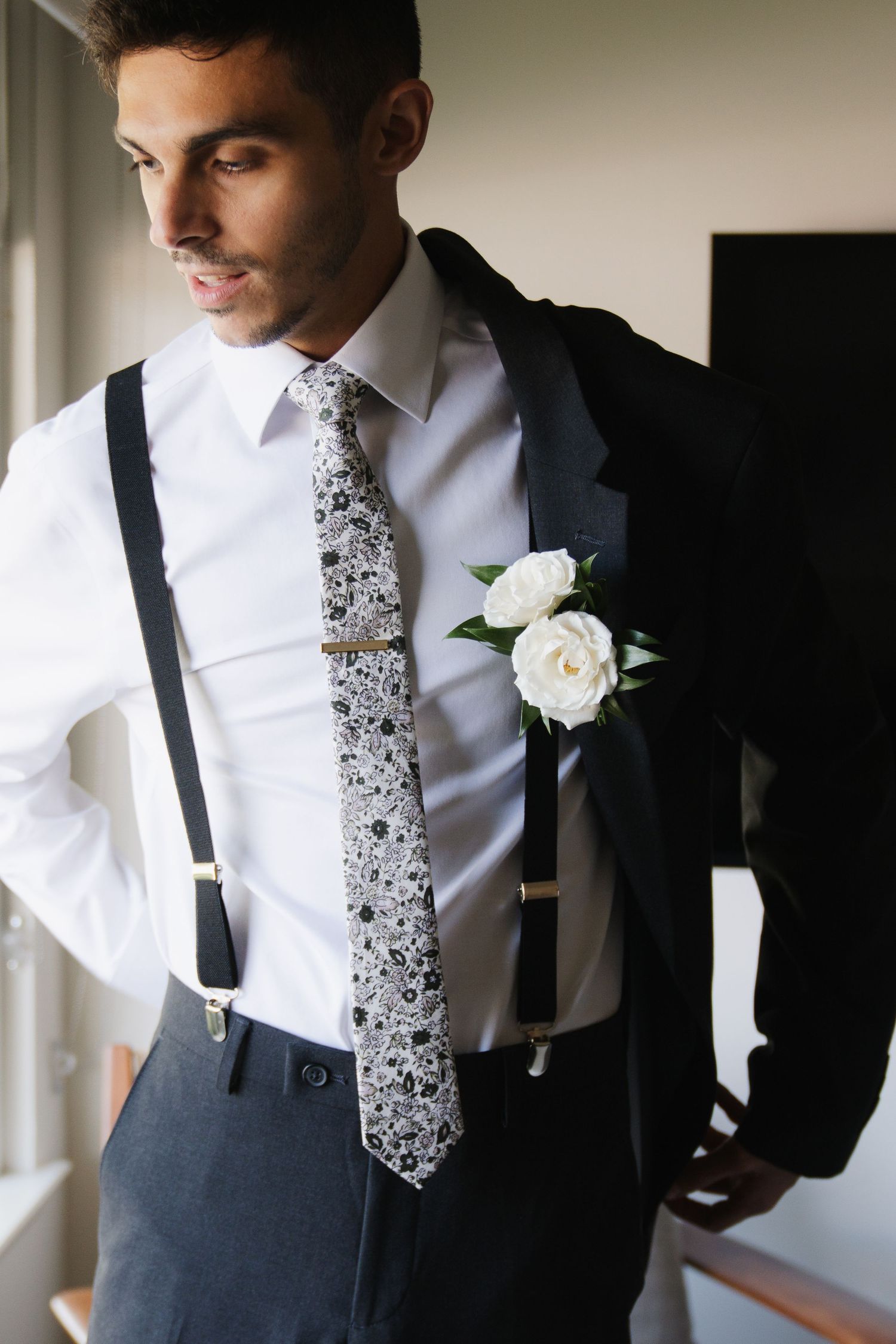 Close-up details of a groom's formal attire showing a white shirt, patterned tie, and black suspenders with a boutonniere.