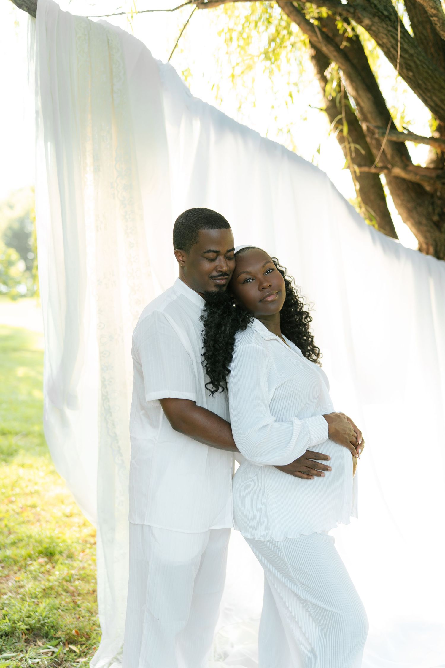 A couple in matching white outfits poses outdoors, the pregnant woman leaning back into her partner's embrace near white draping.