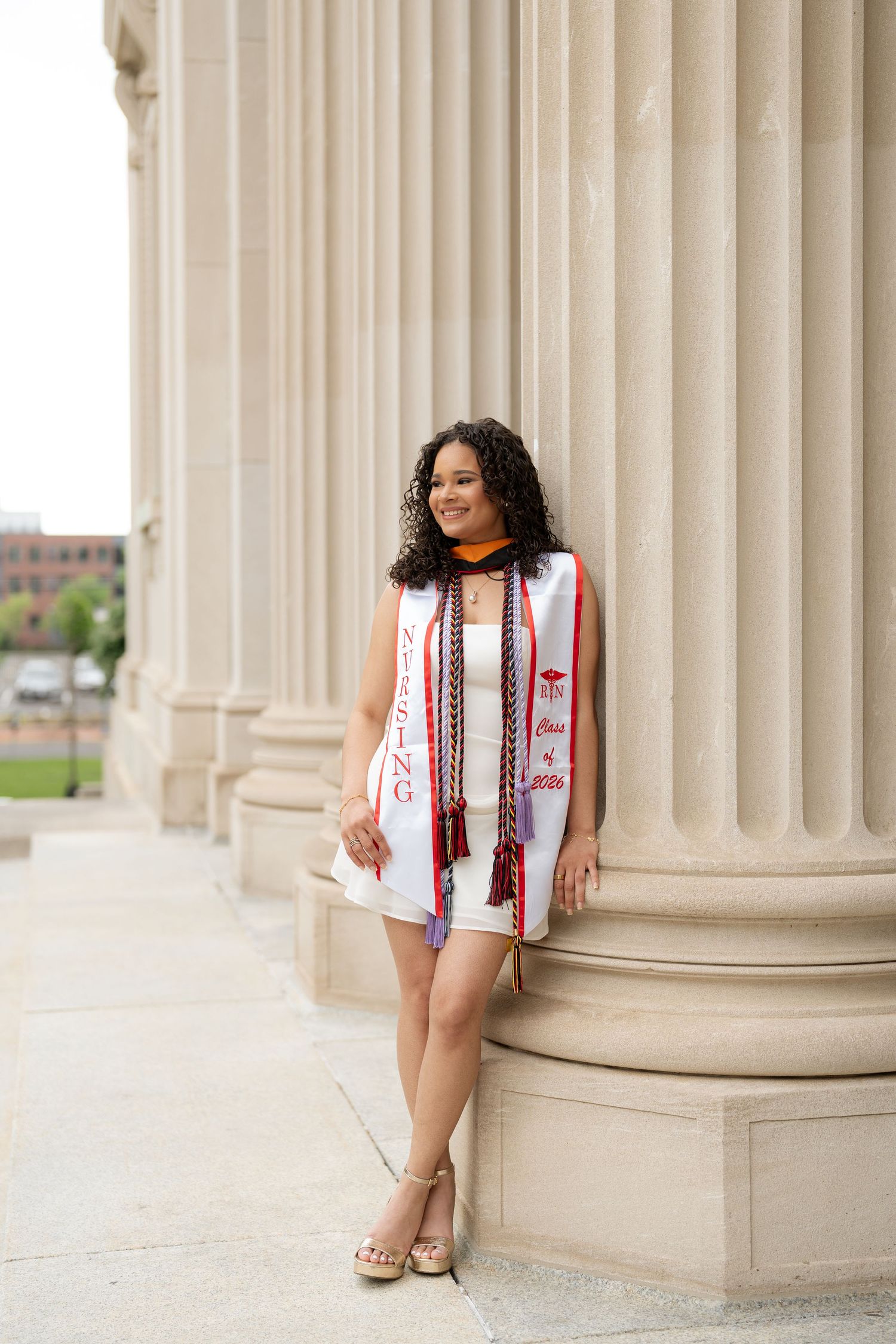 Nursing graduate poses in white dress and graduation stole beside stone columns.