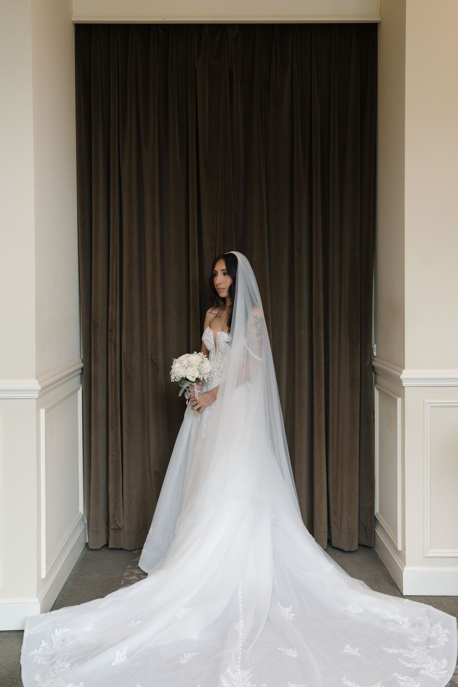 Bride in white lace gown with long veil holds bouquet before dark curtain backdrop.