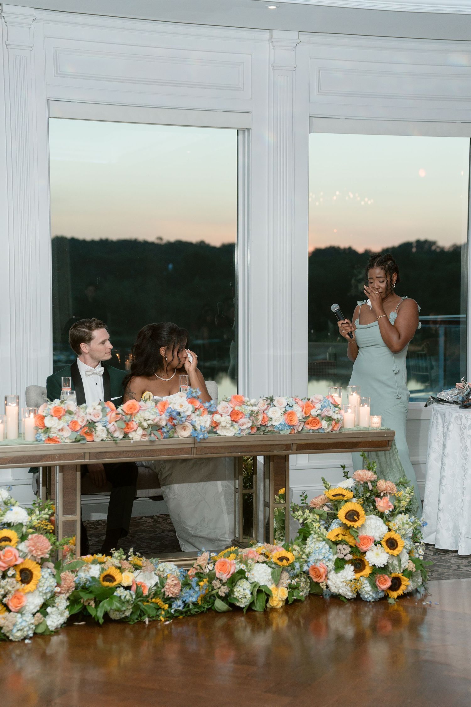 Maid of honor gives a heartfelt speech at sweetheart table adorned with sunflowers at sunset.