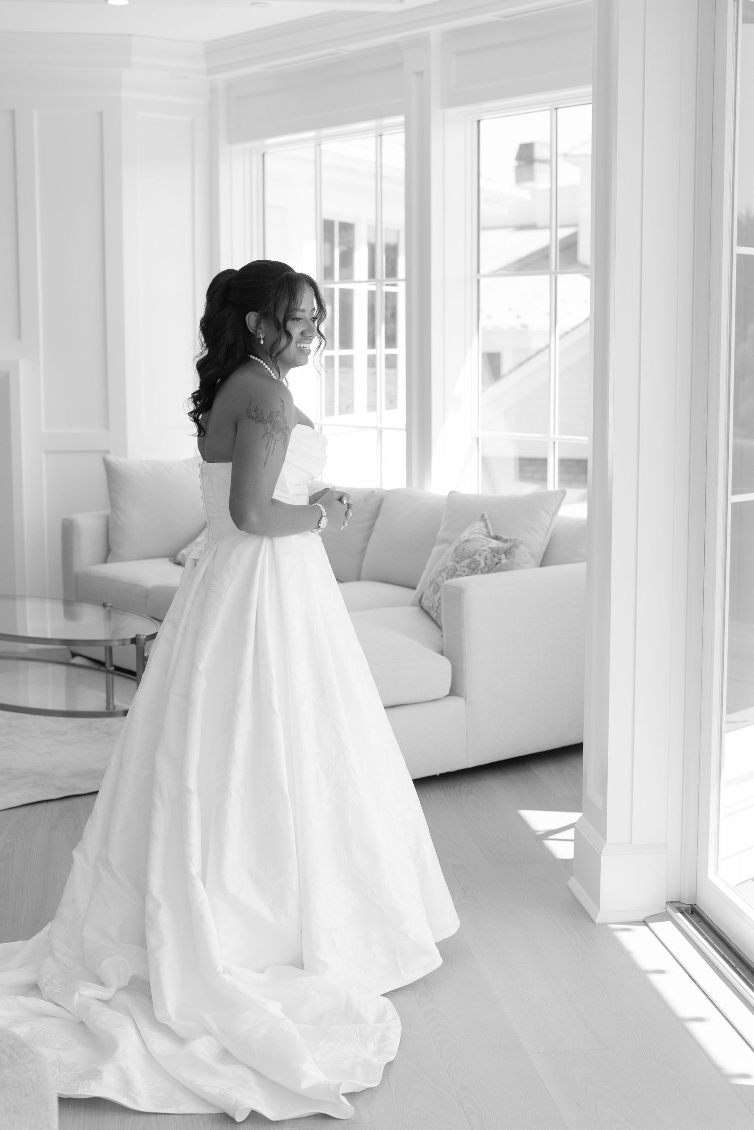 Black and white photo of a bride in a strapless ball gown standing by bright windows in an elegant white room.