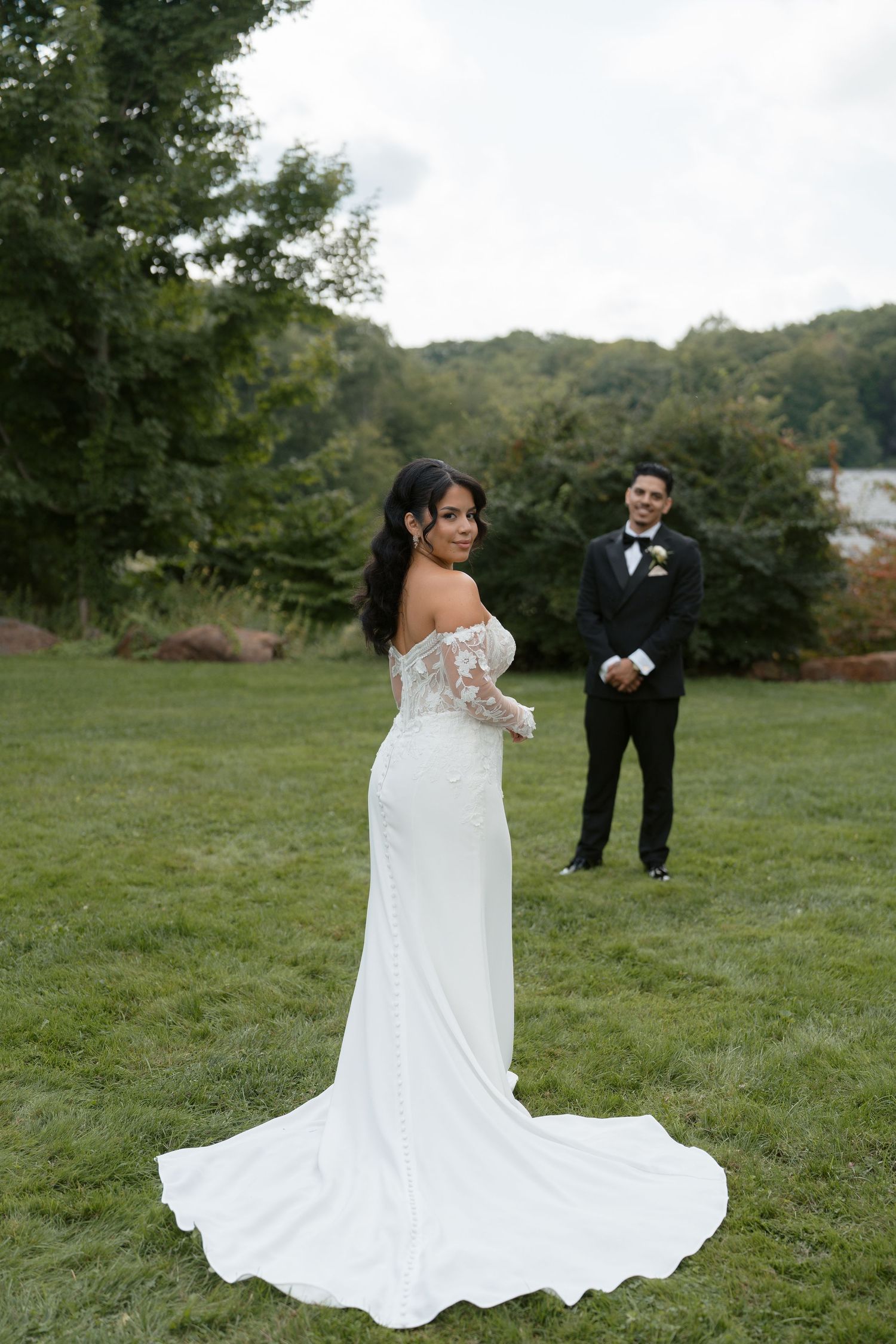 Bride in off-shoulder white gown with train poses outdoors while groom in black tuxedo stands behind her.