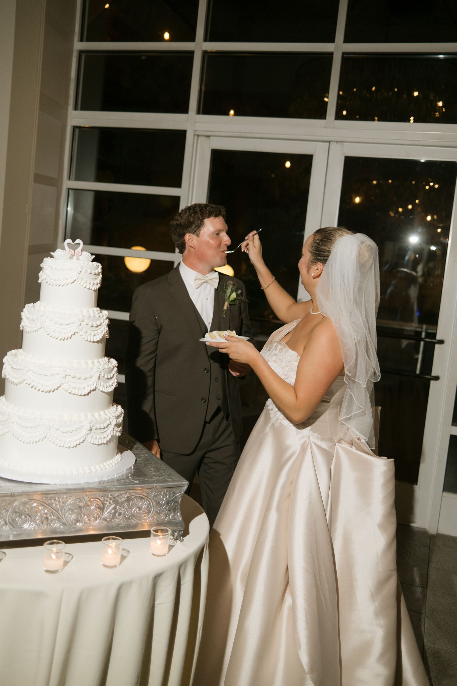 Bride feeds wedding cake to groom beside elegant four-tier white cake at night reception.