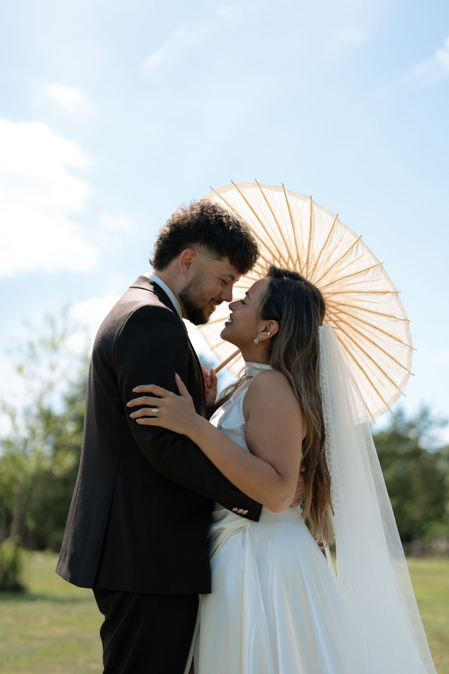 Bride and groom share a romantic moment under a parasol umbrella against a blue sky backdrop.