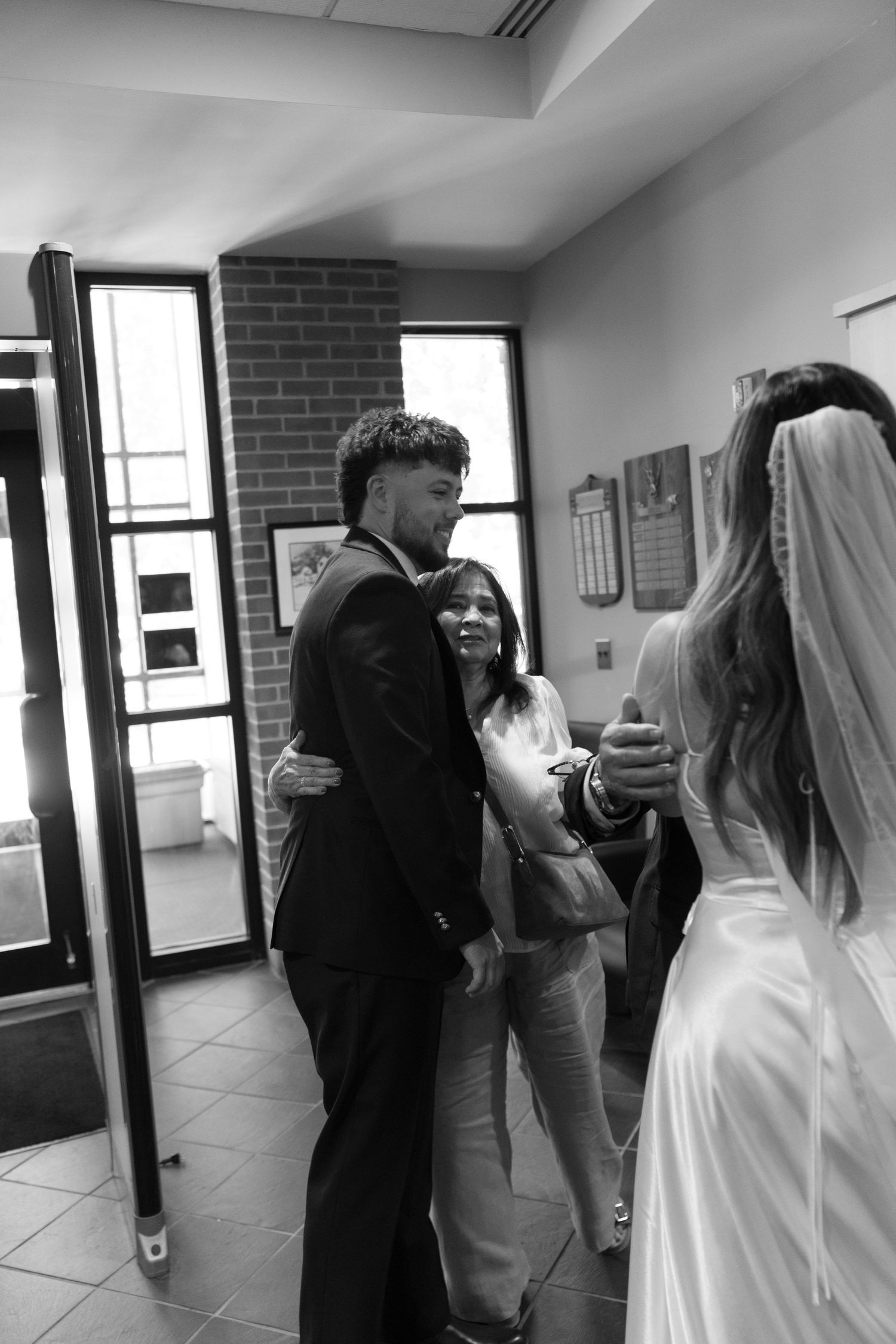 Black and white photo of groom embracing guest while bride stands nearby in wedding venue.
