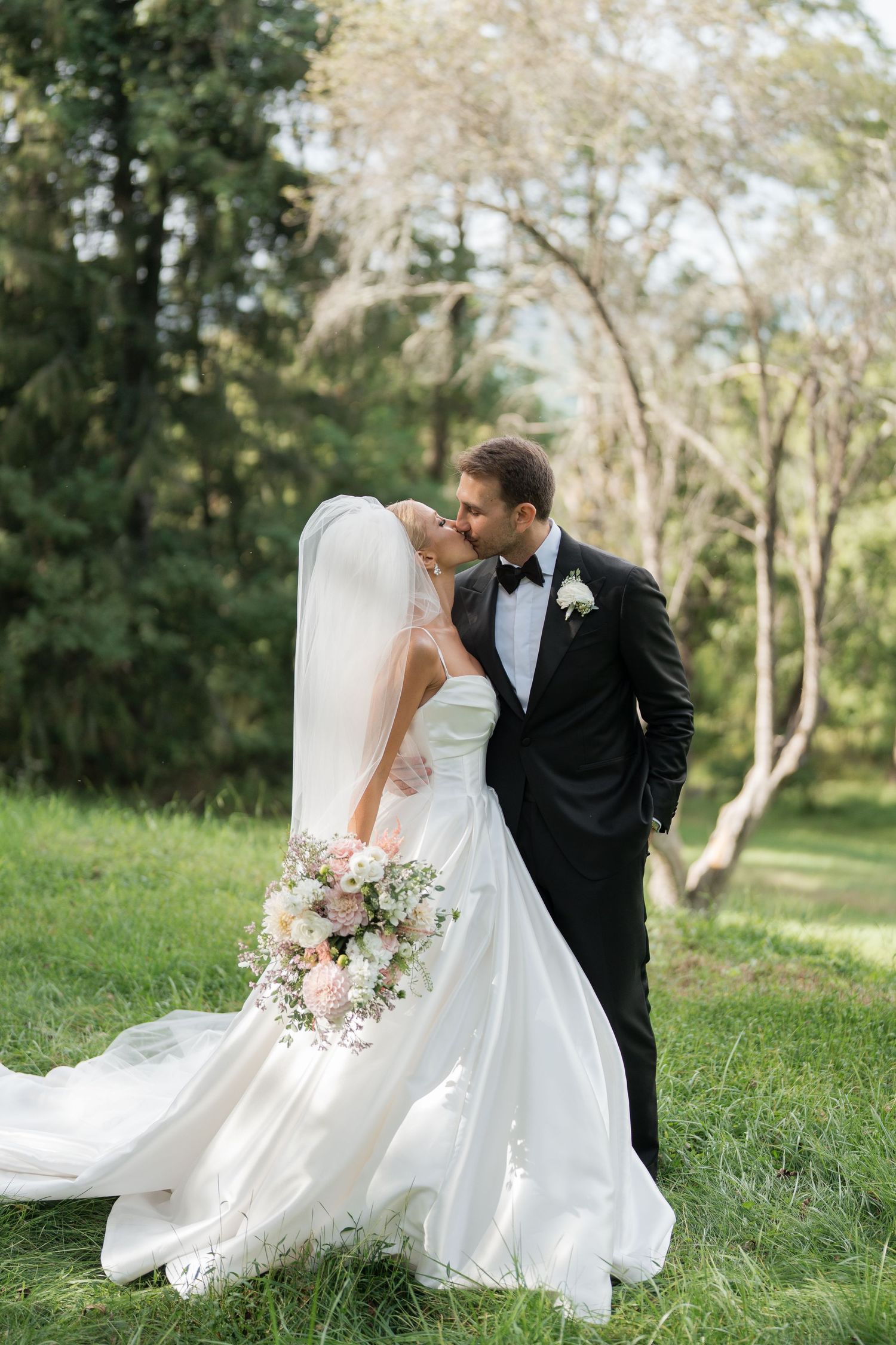 A bride and groom share a romantic kiss outdoors, surrounded by lush greenery, as her white gown and veil flow gracefully.