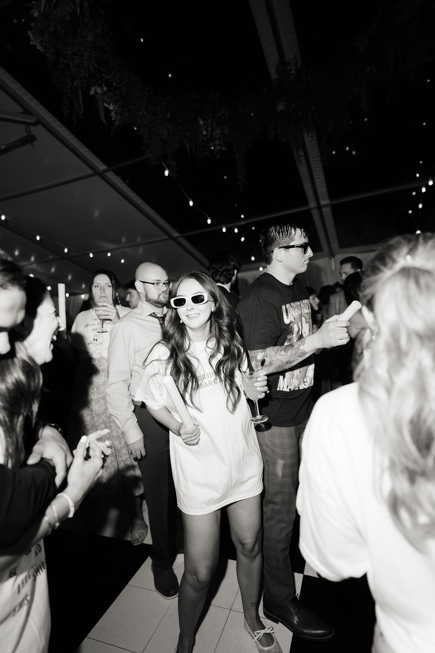 A group of people dancing energetically at a party under string lights in black and white photography.