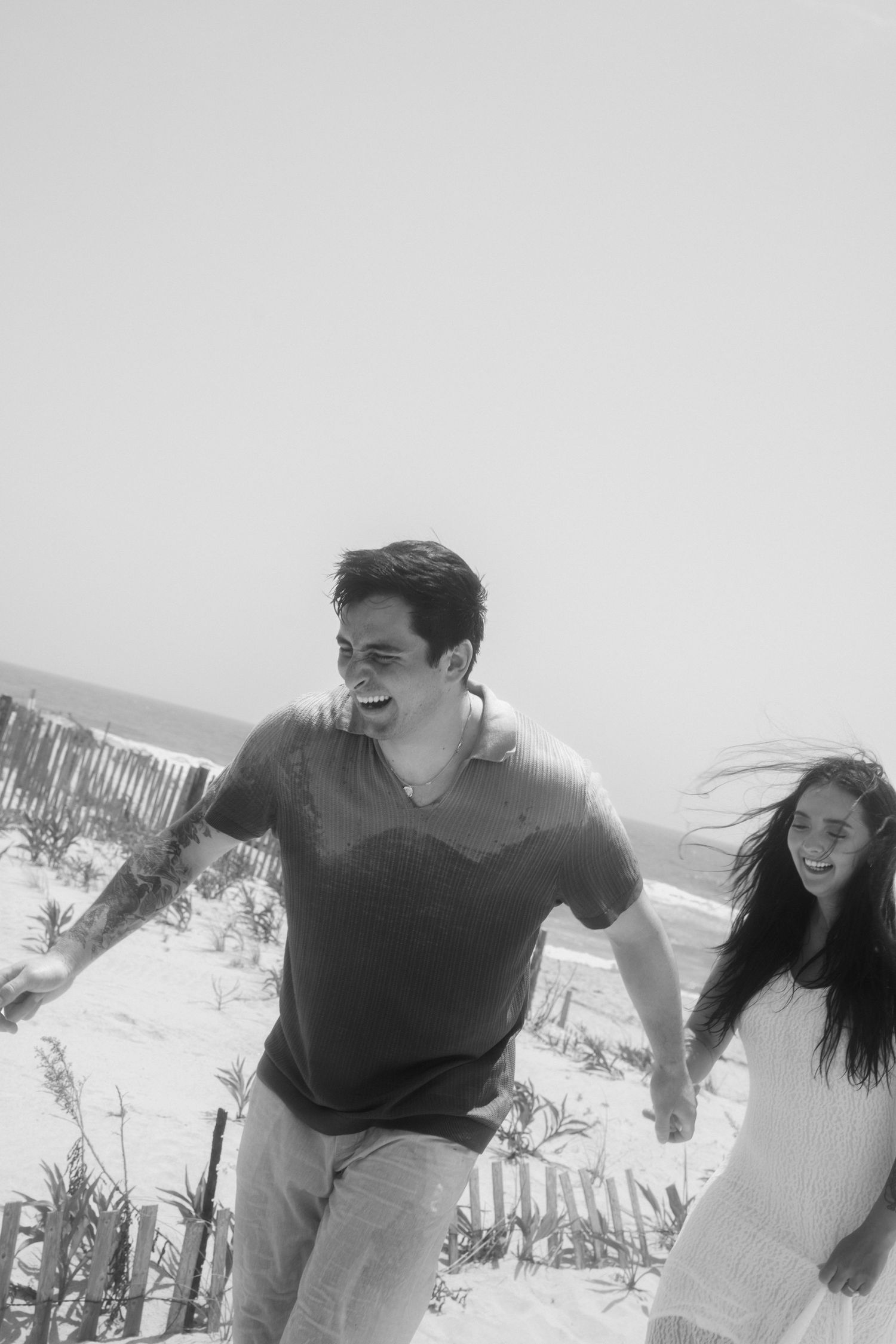 A couple runs joyfully along a sandy beach path with wooden fences on a bright sunny day.