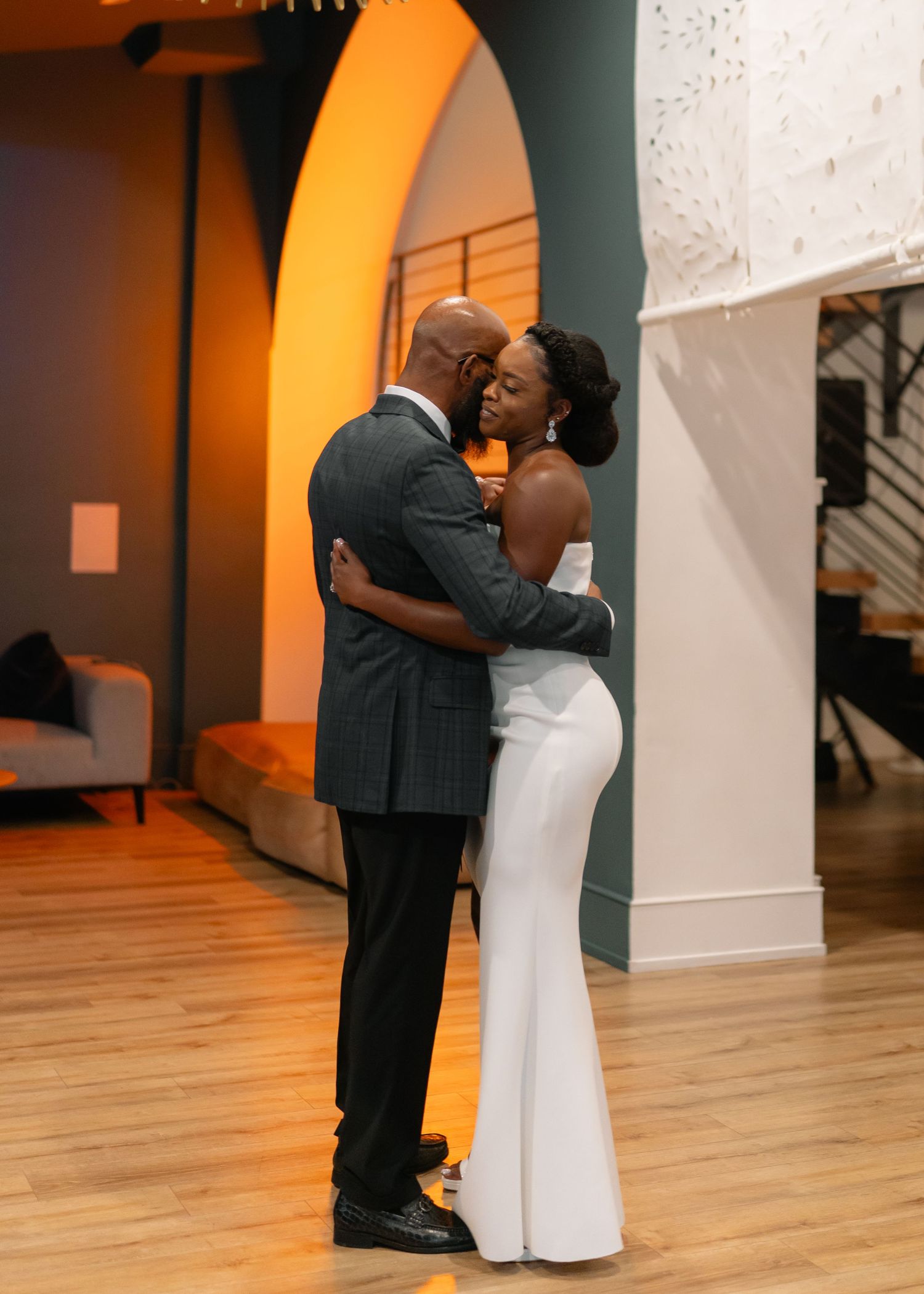 Dancers move gracefully across a wooden floor under arched ceilings with warm orange lighting.