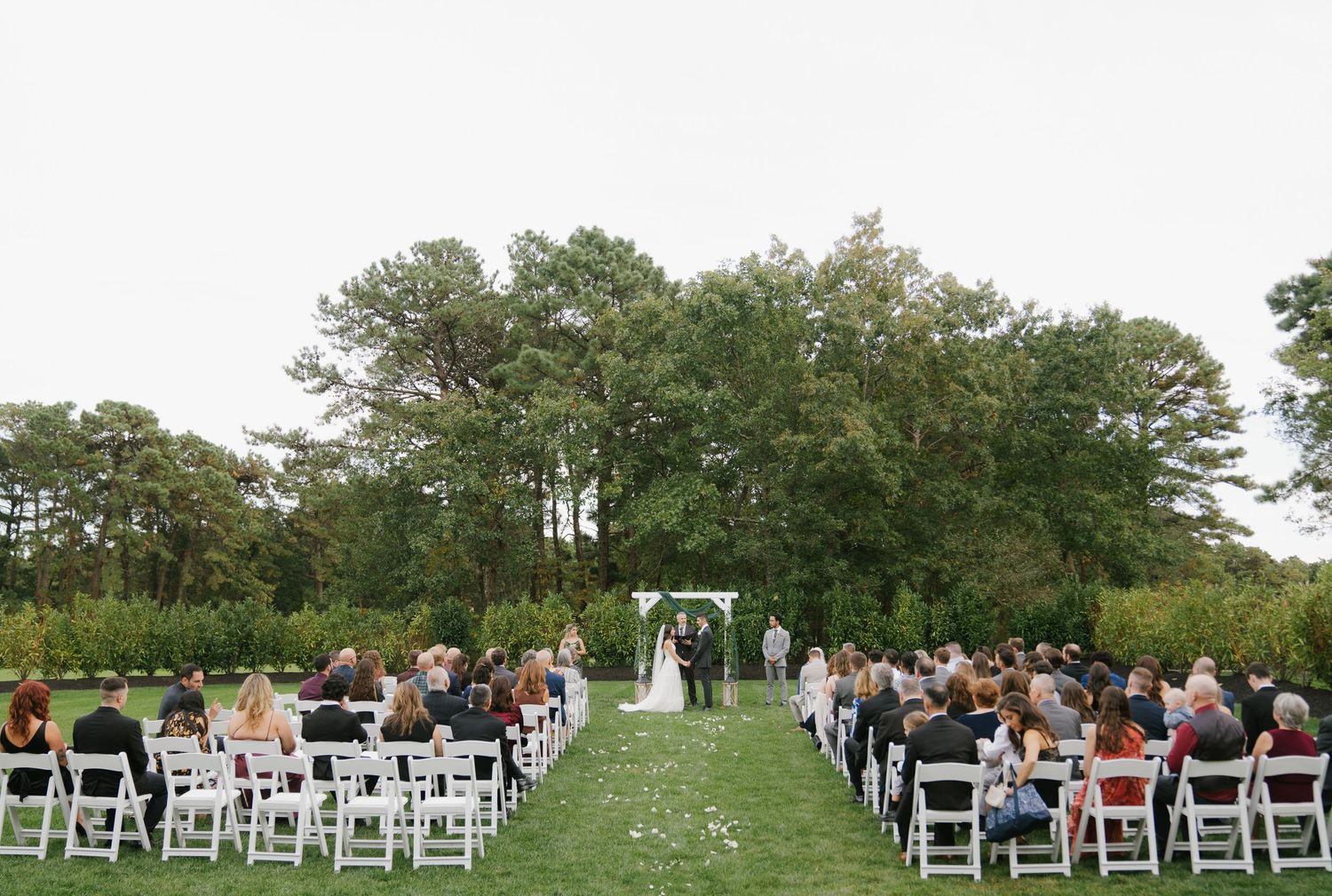 Outdoor wedding ceremony with white chairs set up on a green lawn surrounded by tall trees in the background.
