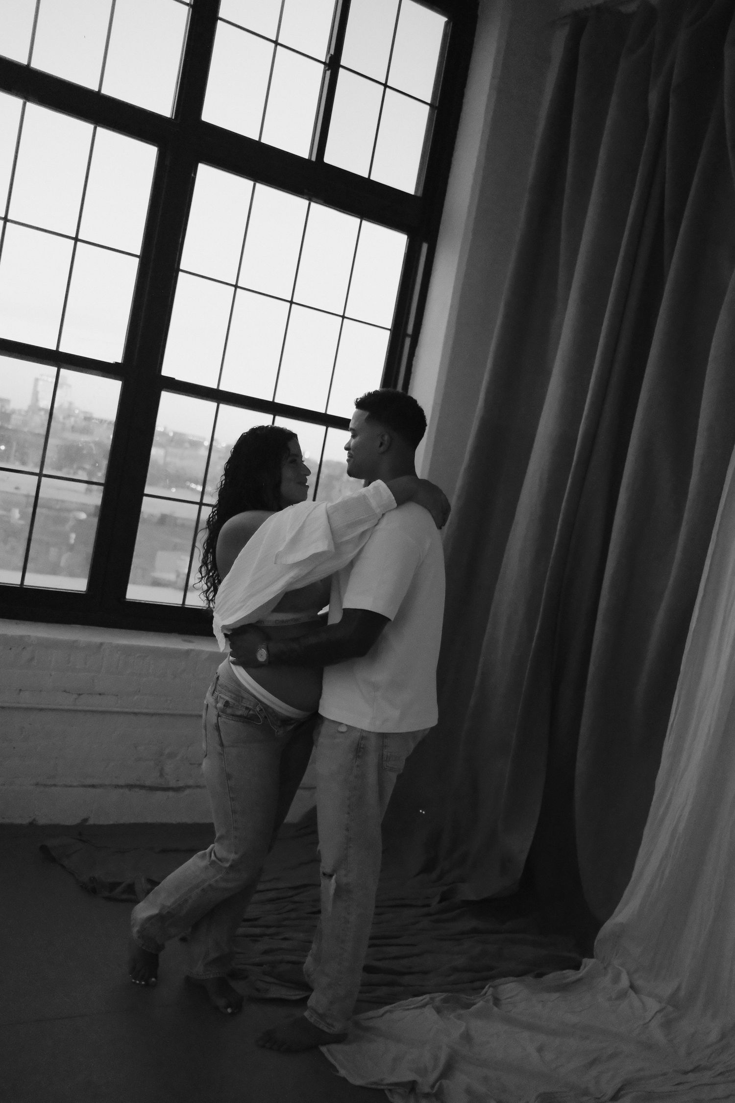 Couple embracing near large industrial window in black and white romantic portrait photo.