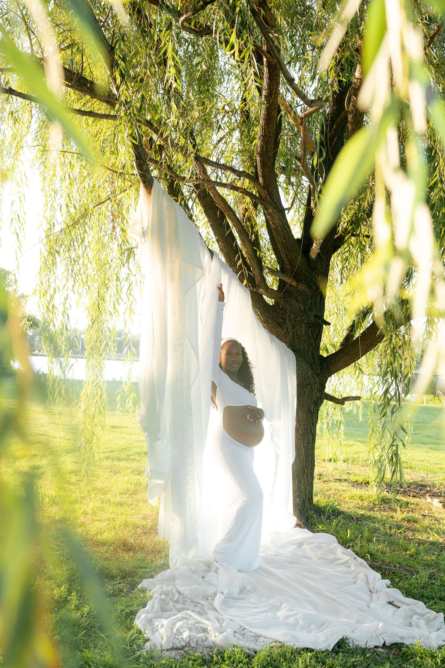 A pregnant woman in white draped fabric posing under a sunlit willow tree outdoors.