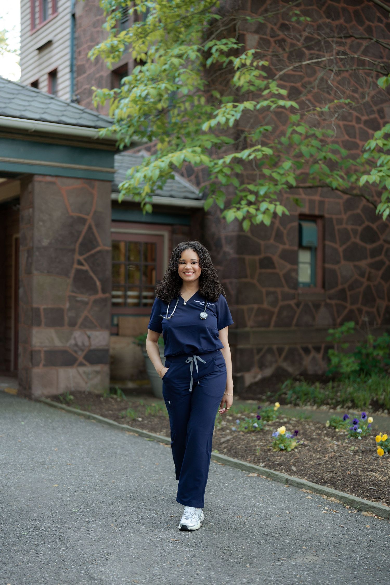 Smiling nurse in navy scrubs with stethoscope walks outside historic stone building.