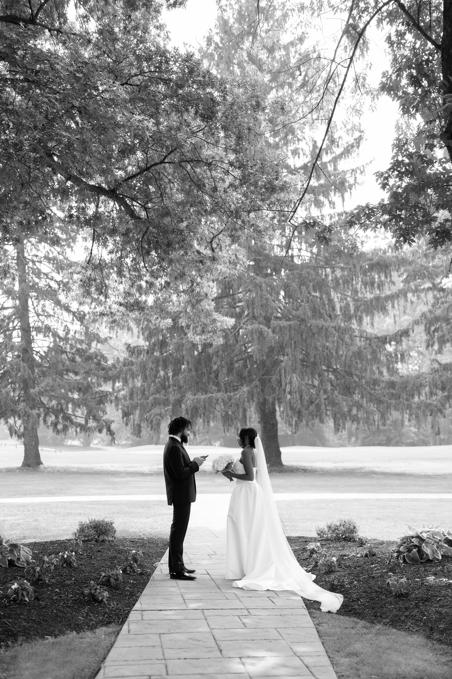 Black and white photo of bride and groom exchanging vows outdoors on a stone path surrounded by tall trees.