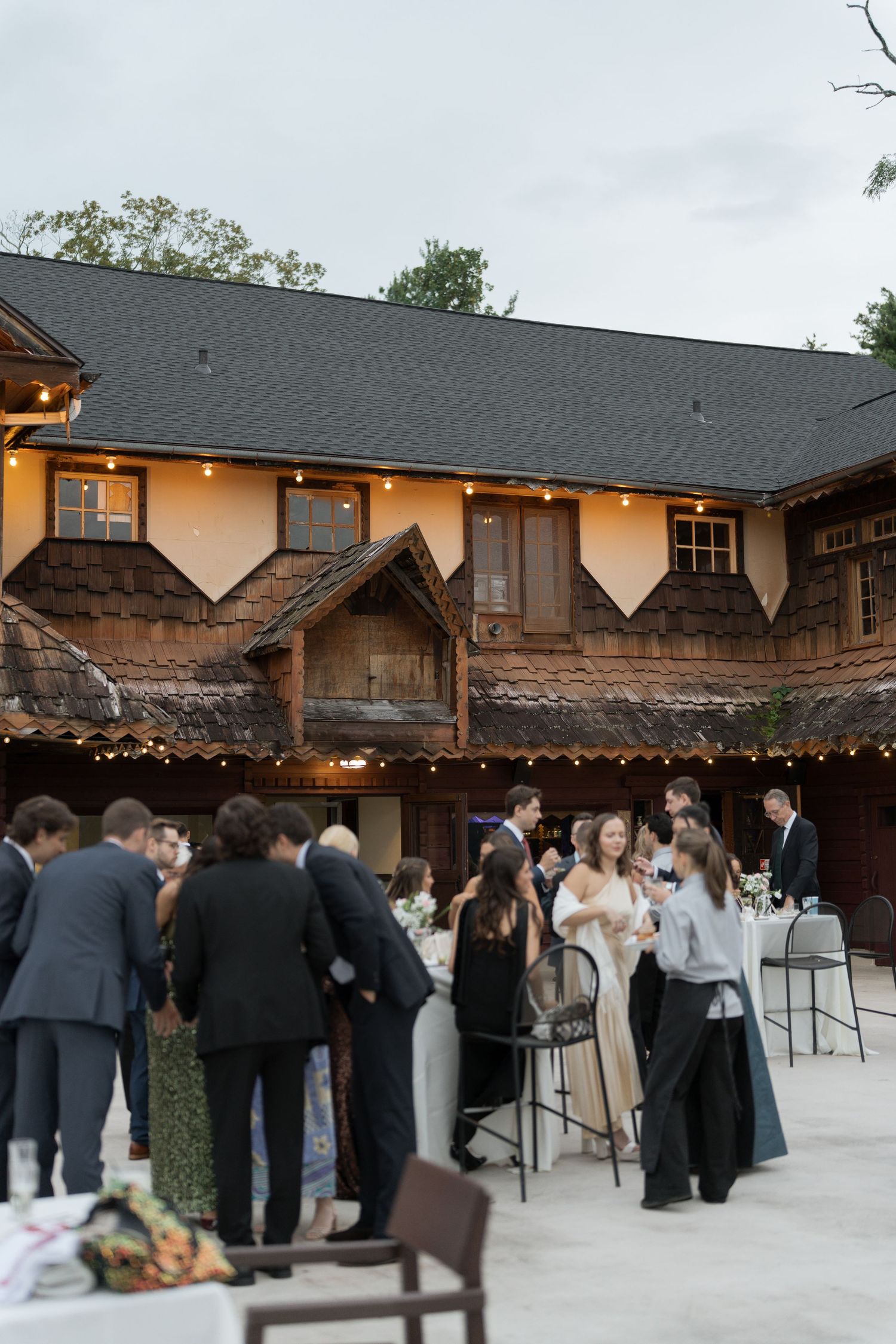 Wedding reception guests mingle outdoors at rustic venue with string lights and wooded surroundings.