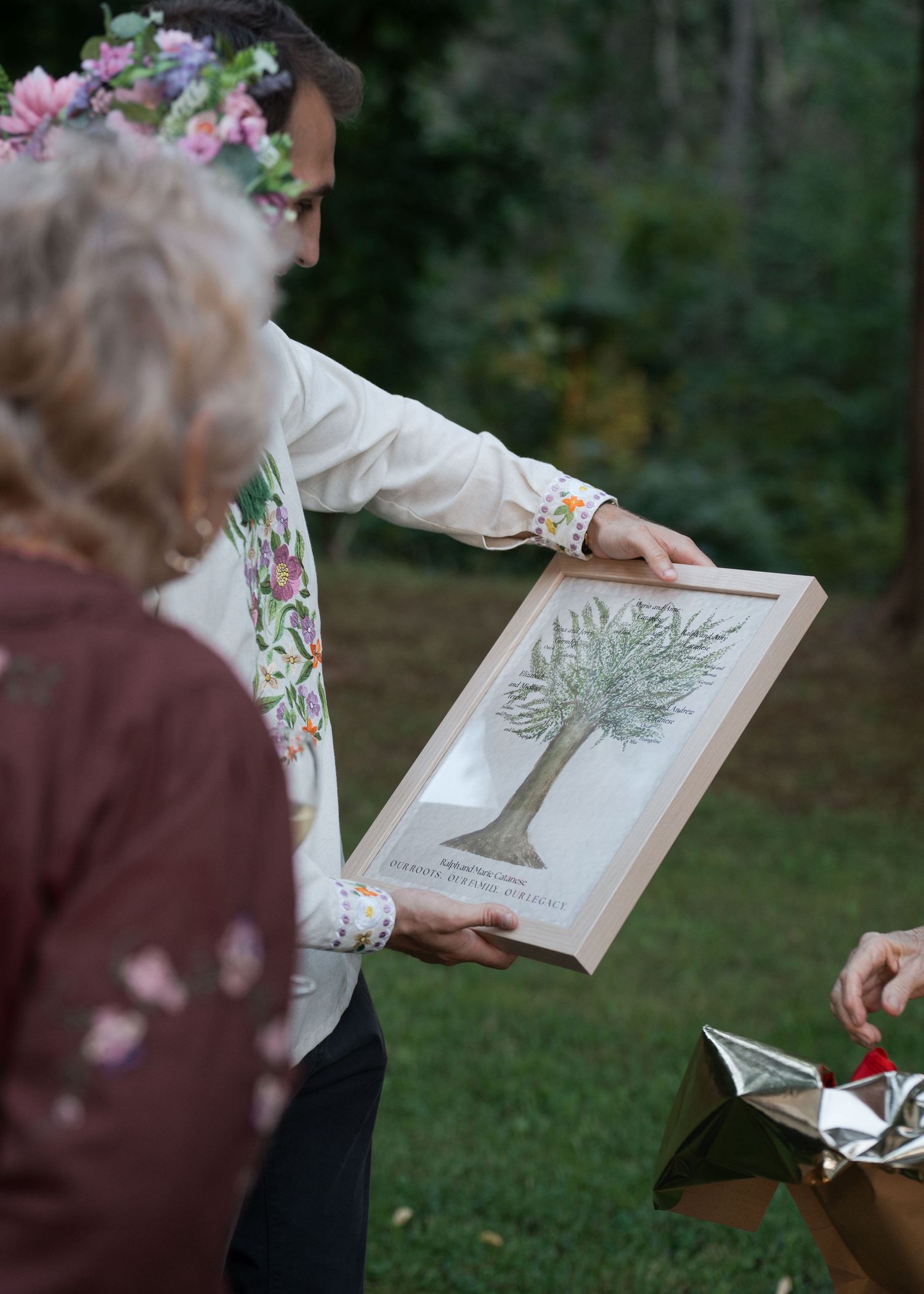 Person in floral crown holding framed tree artwork at outdoor wedding ceremony gift exchange.