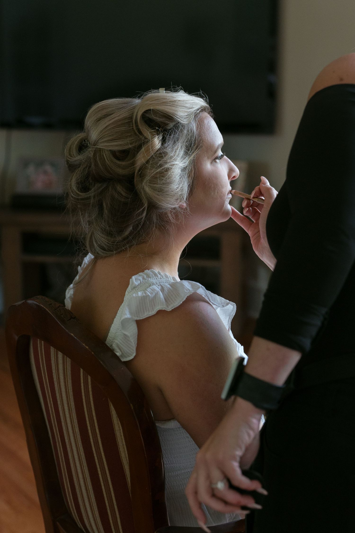 Bride getting makeup applied by a makeup artist while seated in a wooden chair in her white wedding dress.