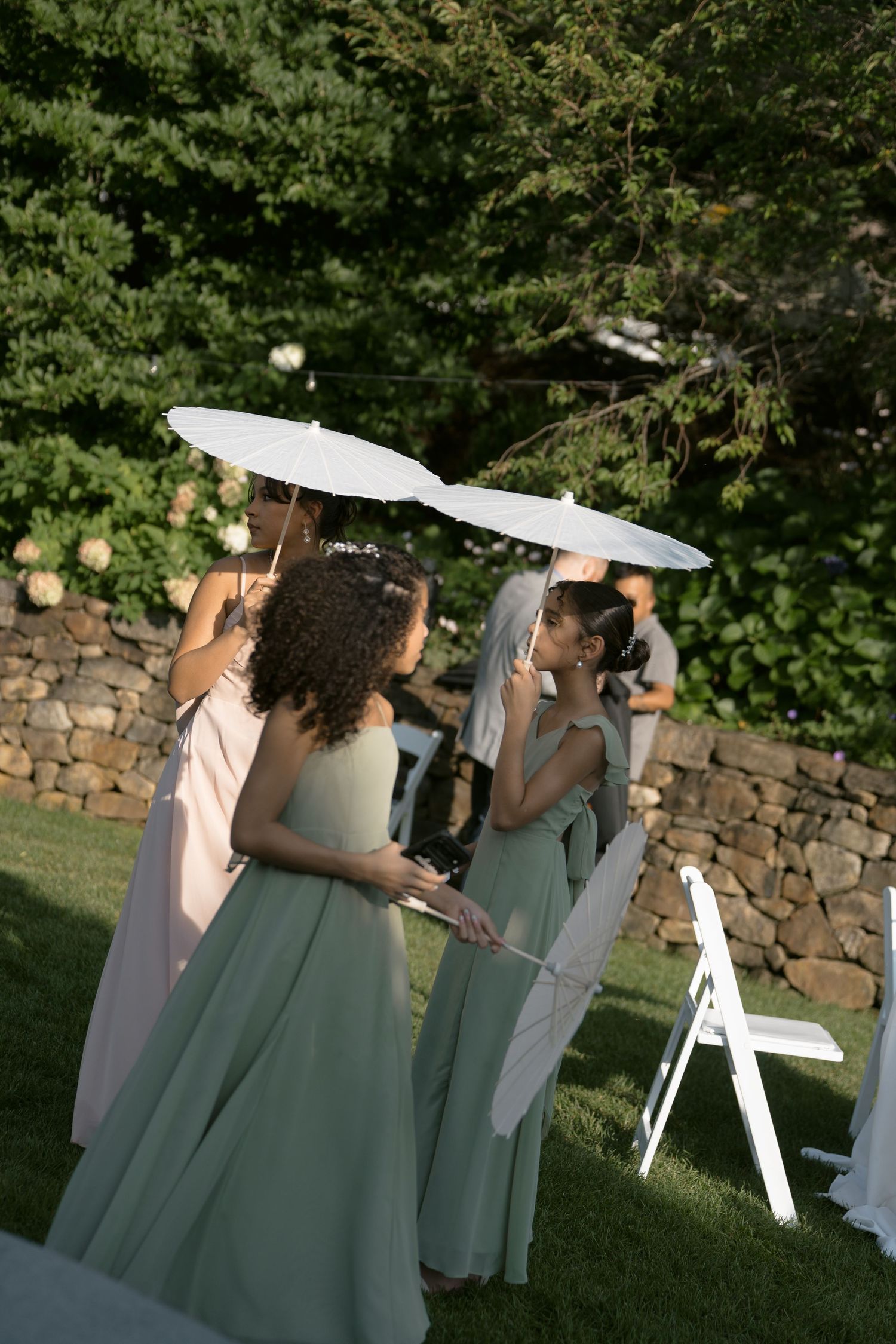 Bridesmaids in sage green gowns holding white parasols during an outdoor wedding ceremony.