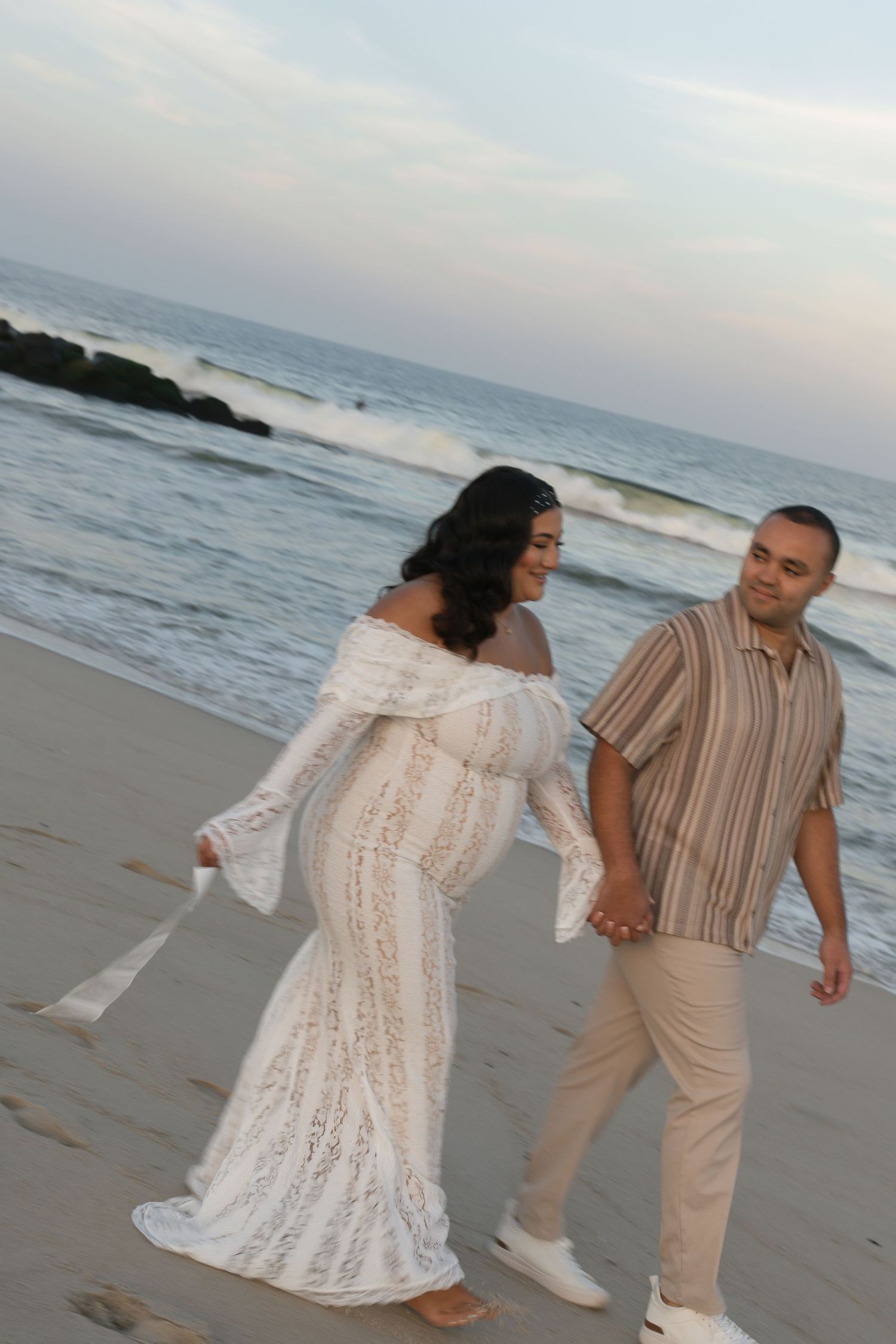 Pregnant woman in white lace maternity gown holds hands with man in striped shirt while walking on beach at sunset.