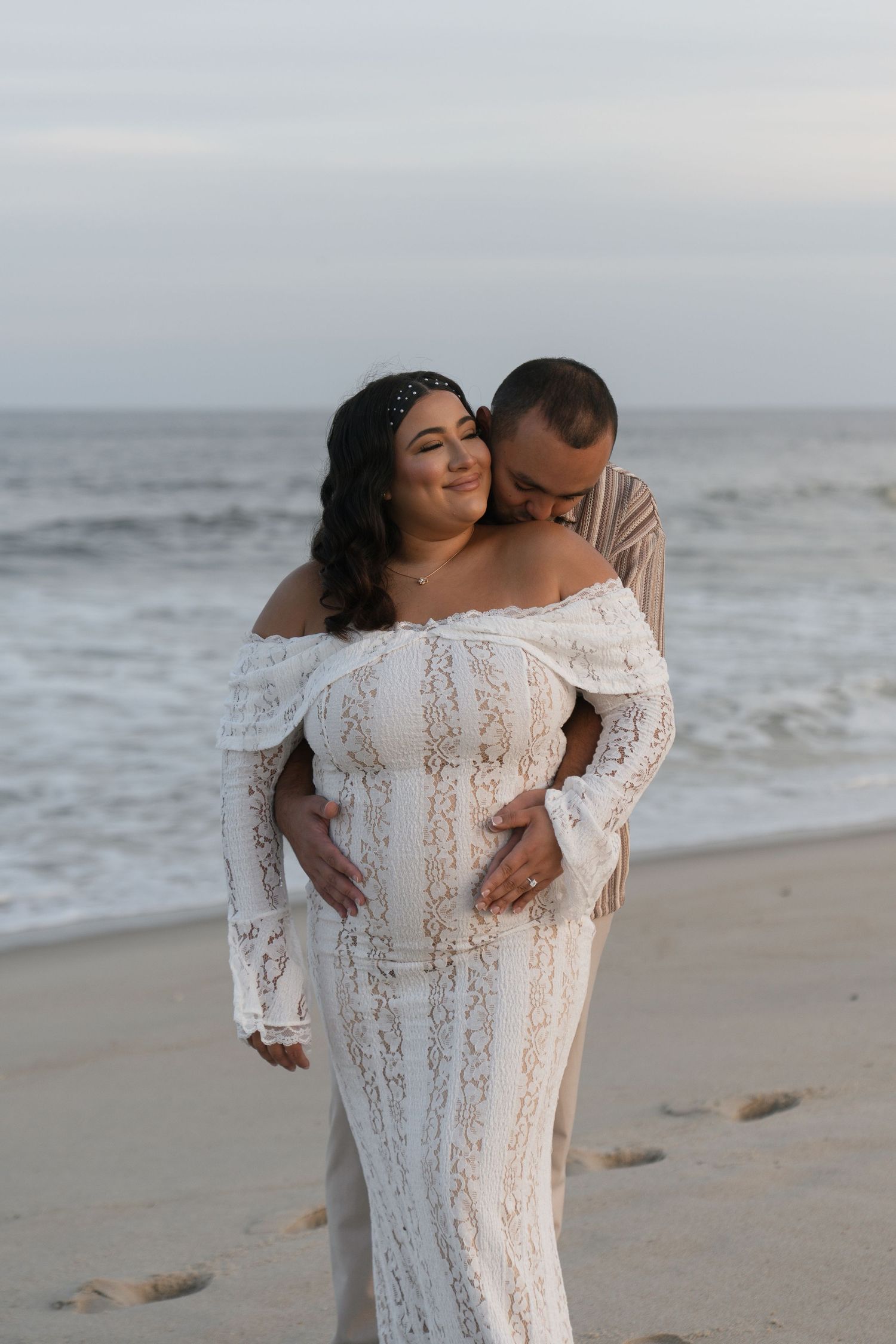 A romantic beach photoshoot featuring a couple in intimate poses wearing white against the ocean backdrop.