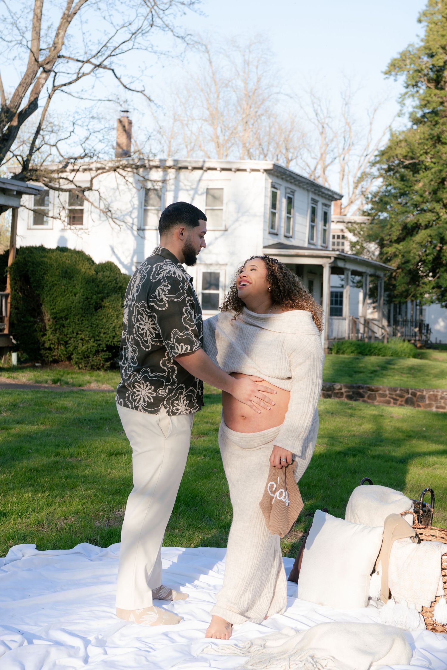 Couple in white outfits share intimate moment on white blanket in outdoor garden setting.
