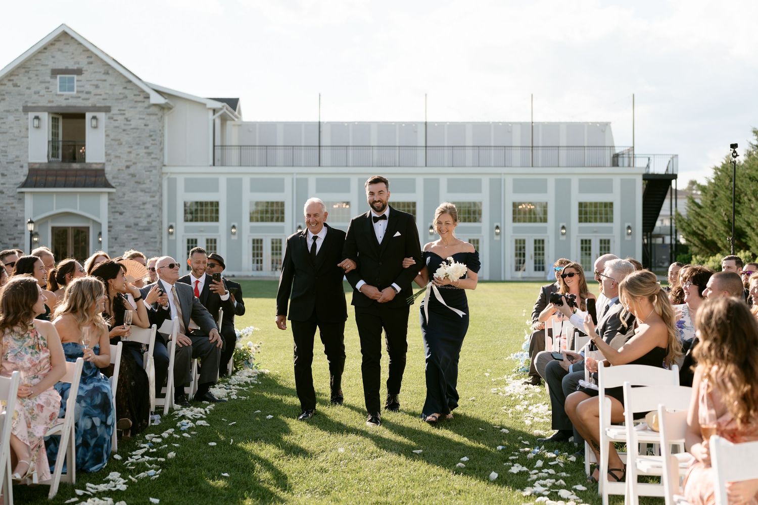 Groom walks down outdoor wedding aisle with family at elegant estate venue on sunny day.