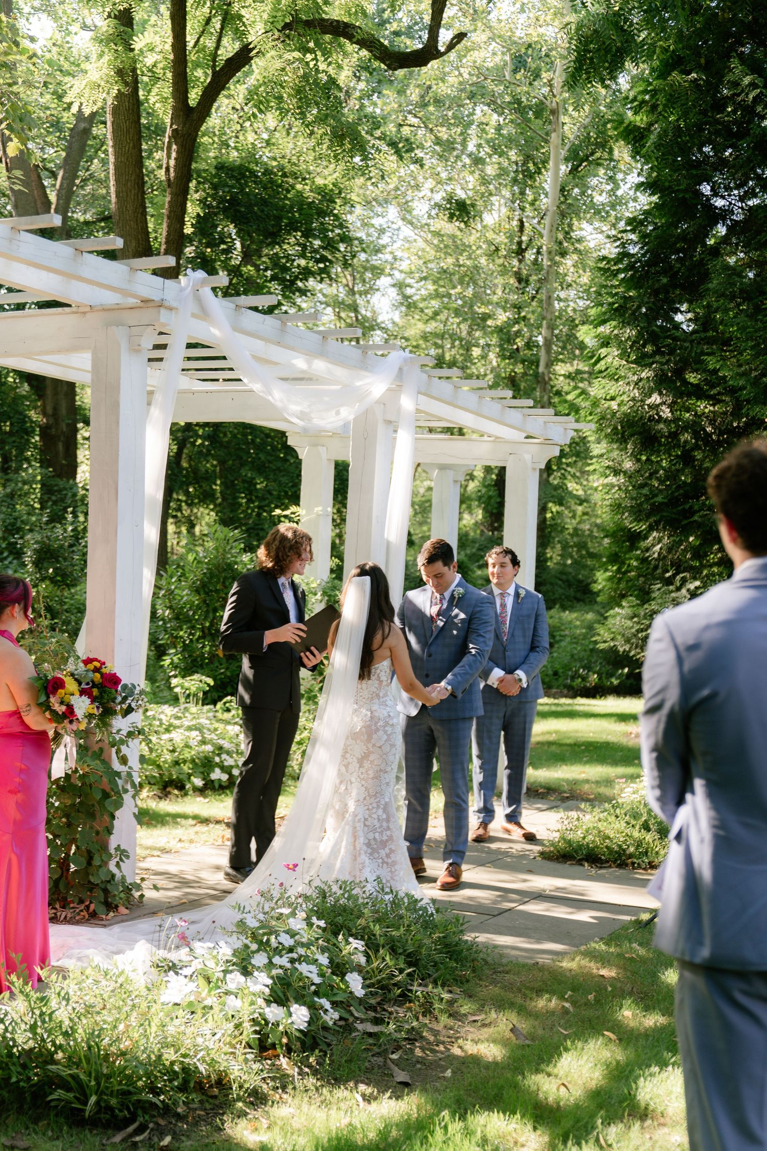 Wedding ceremony taking place under a white pergola surrounded by greenery and flowers.