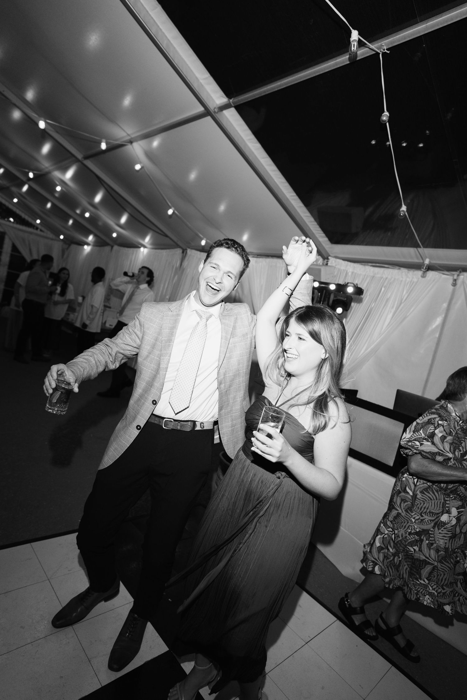 Black and white photo of guests dancing and celebrating at a wedding reception under string lights.