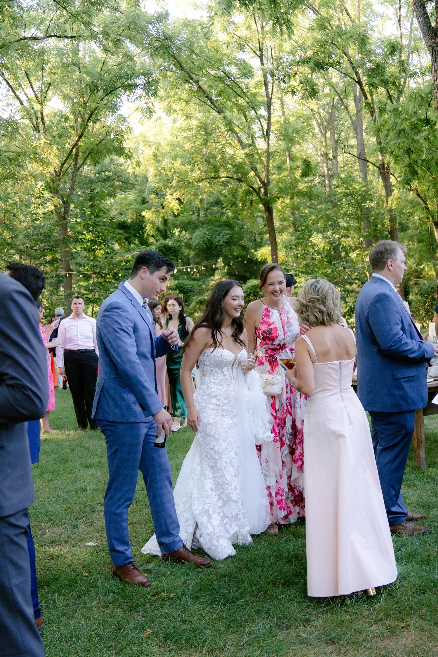 Wedding guests gather outdoors under trees during a summer celebration.