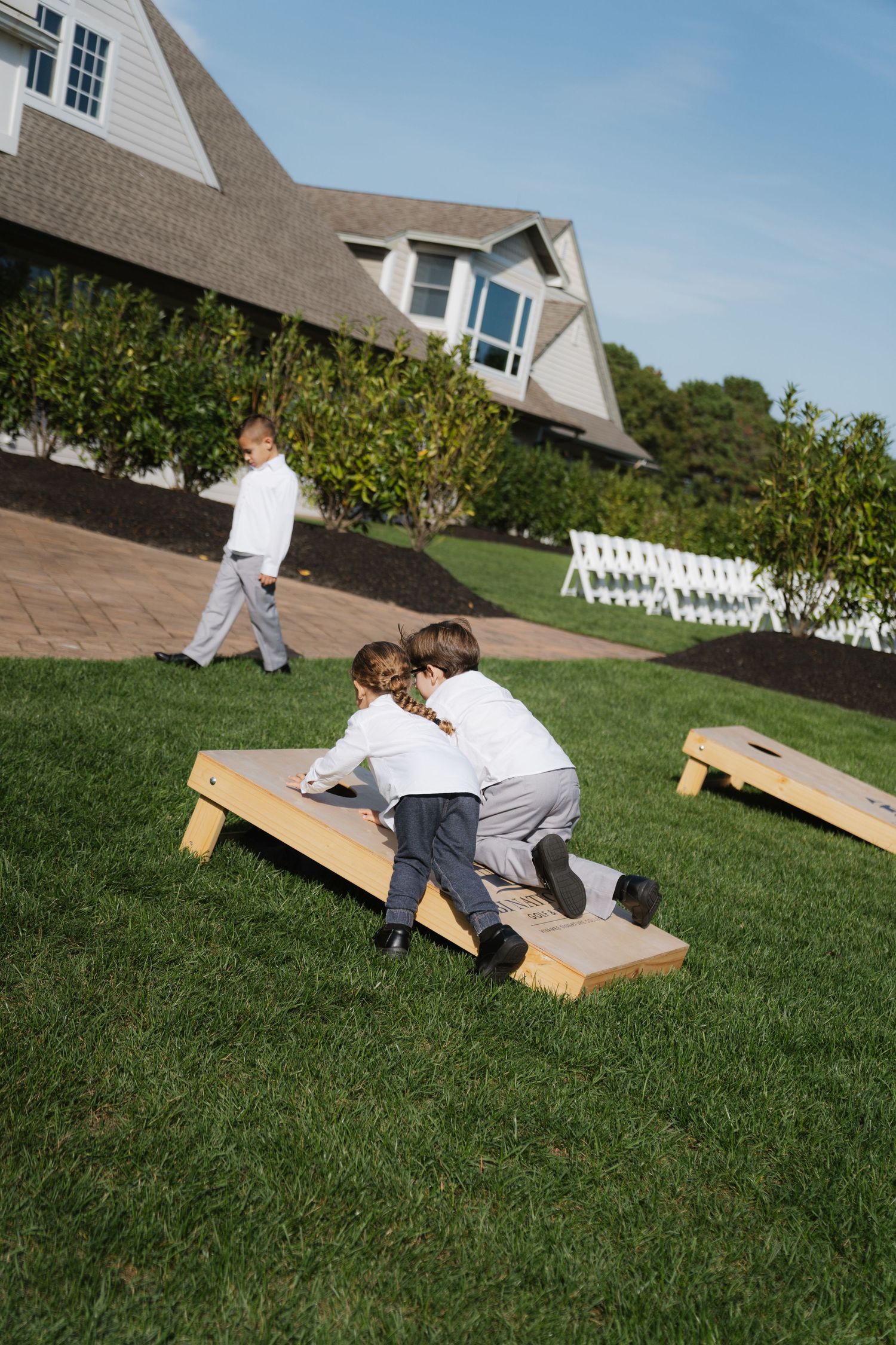 Children playing and sliding down a wooden ramp on a grassy lawn outside a suburban house on a sunny day.
