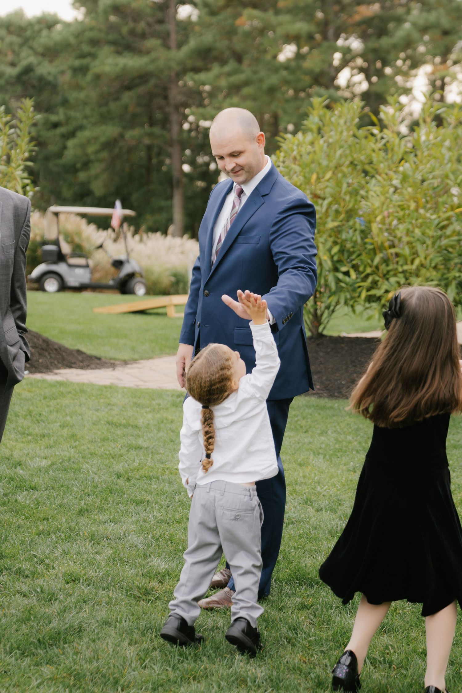 People in formal attire dance and celebrate outdoors on a grassy lawn with trees in the background.