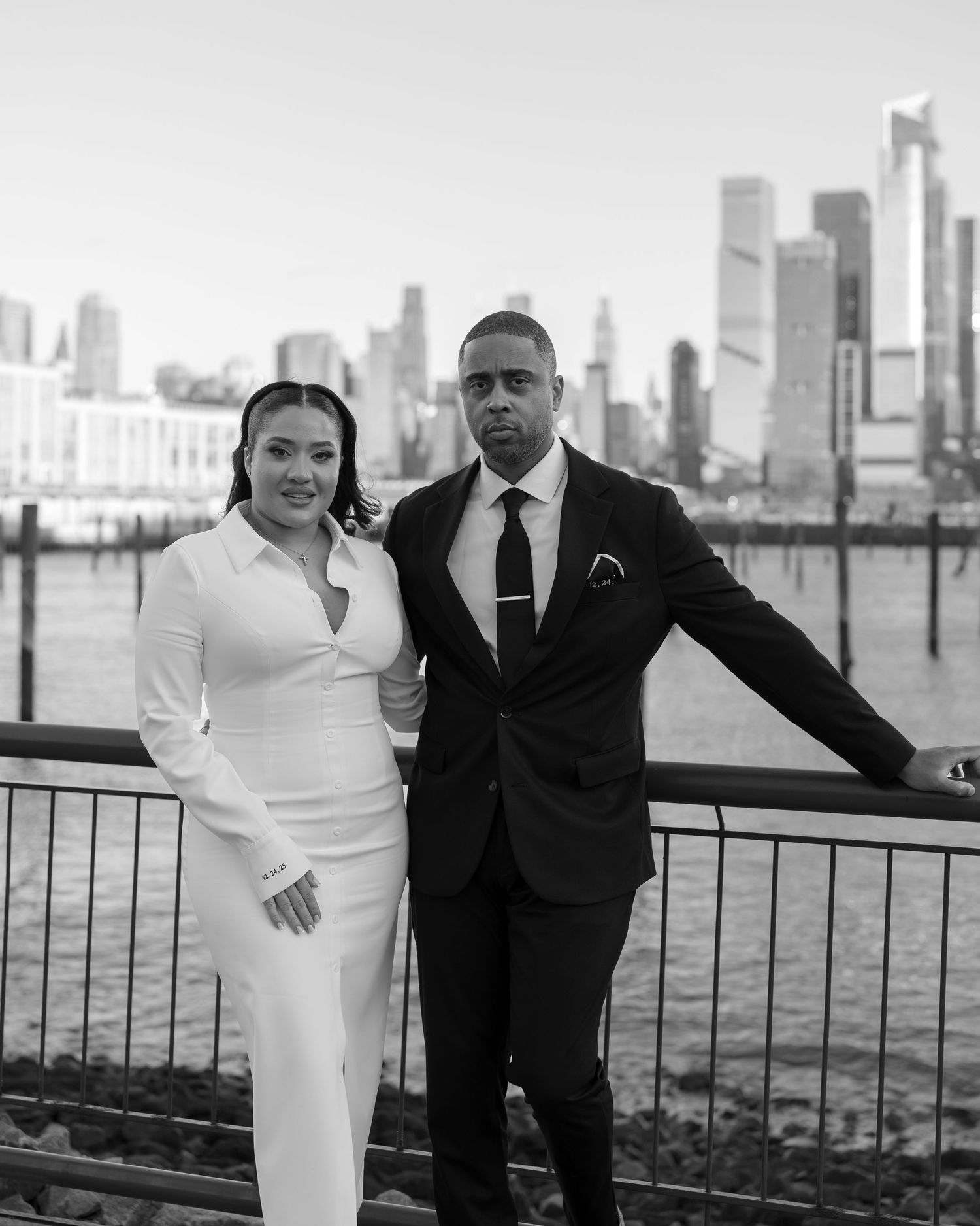 A couple poses against a waterfront railing with a city skyline in the background.