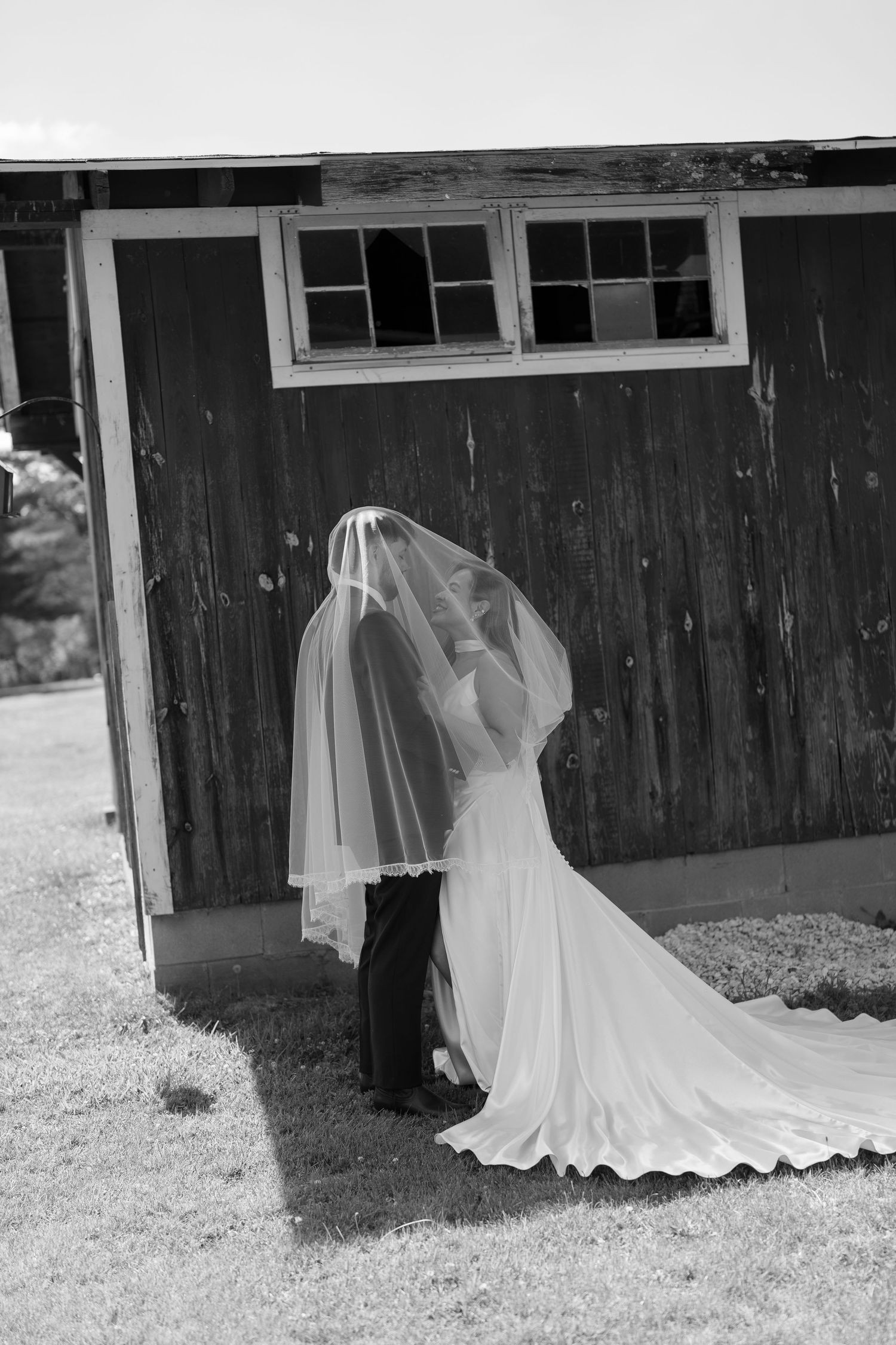 Black and white photo of bride and groom kissing under veil beside rustic wooden barn.