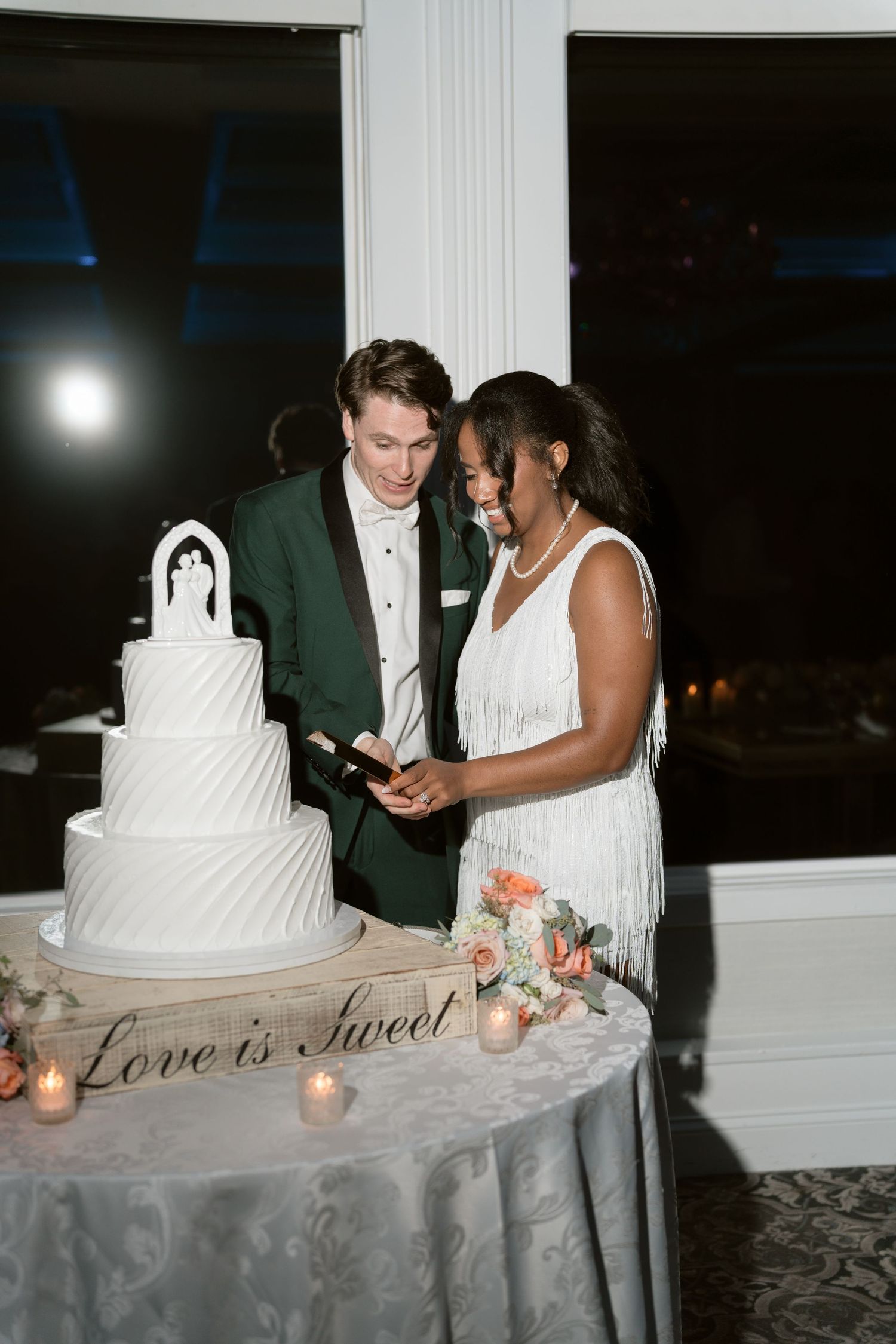 Interracial bride and groom smile while cutting a tiered white wedding cake labeled Love is Sweet.