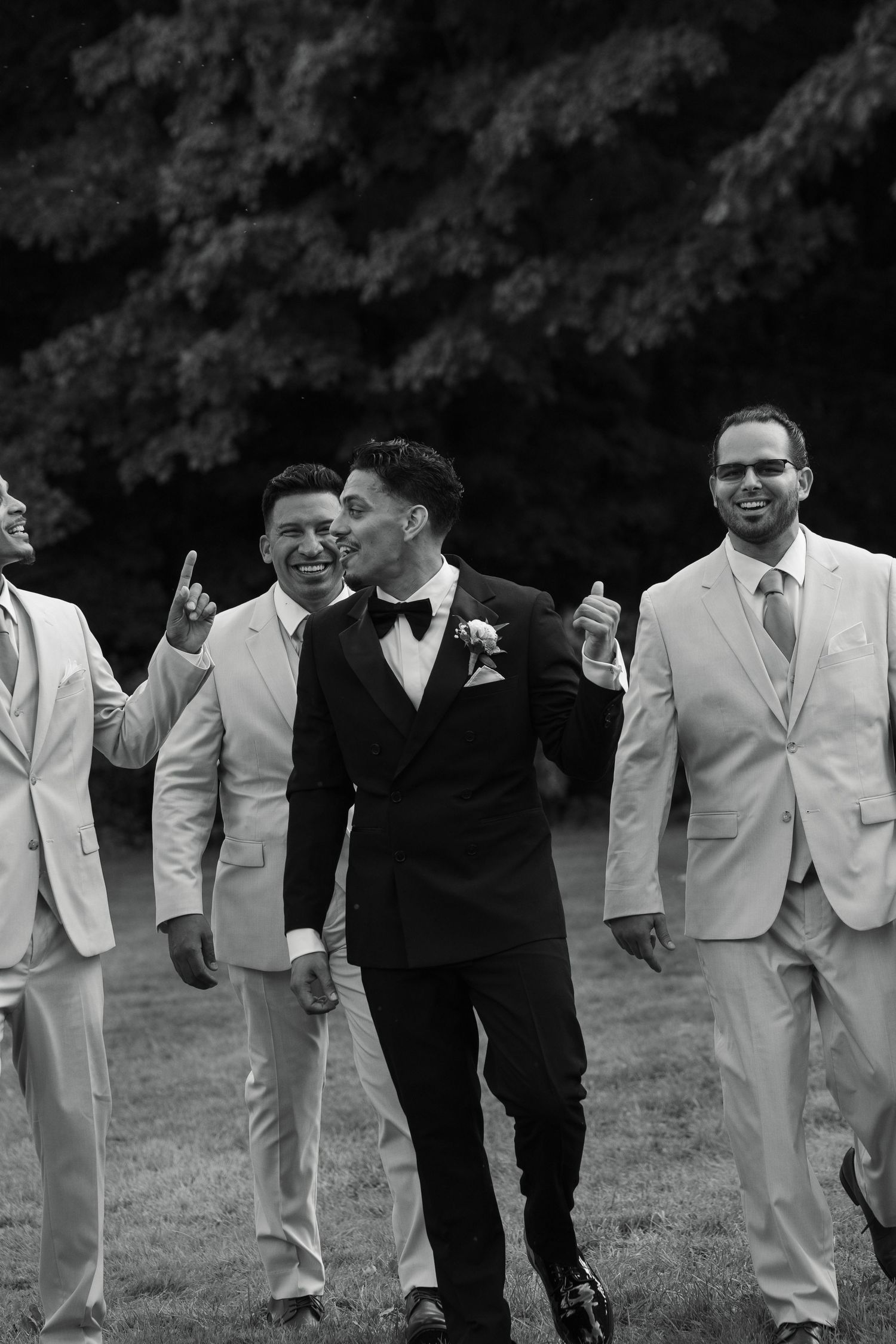 Groom in black tuxedo walks with smiling groomsmen in ivory suits outdoors in black and white wedding photo.