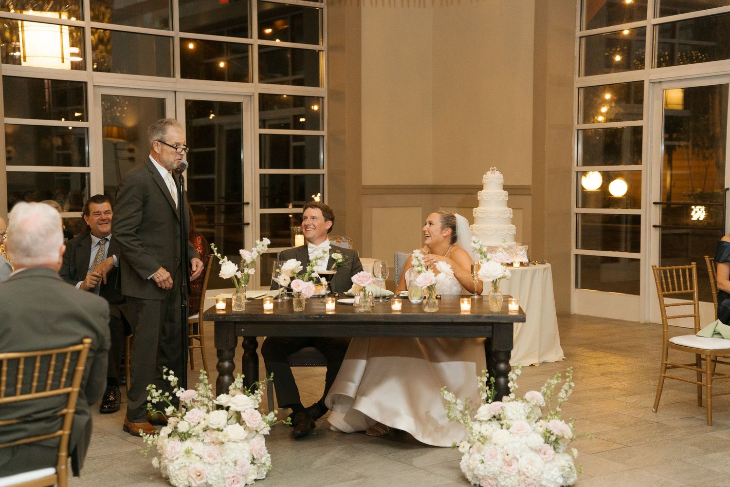 Bride and groom seated at sweetheart table during wedding reception toast with white floral arrangements.
