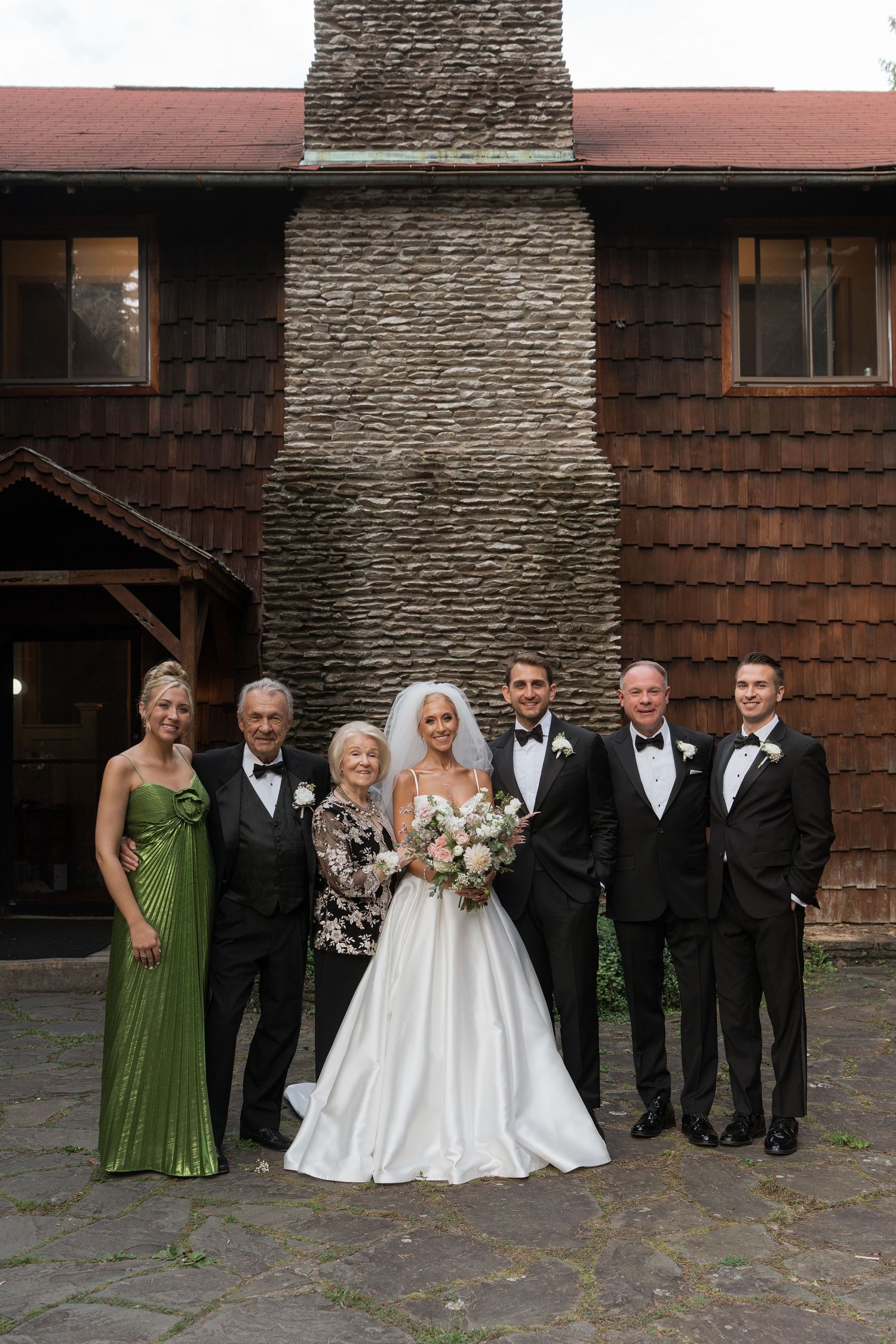 Wedding party poses in front of rustic stone building, bride in white gown with bouquet, groom and groomsmen in tuxedos.