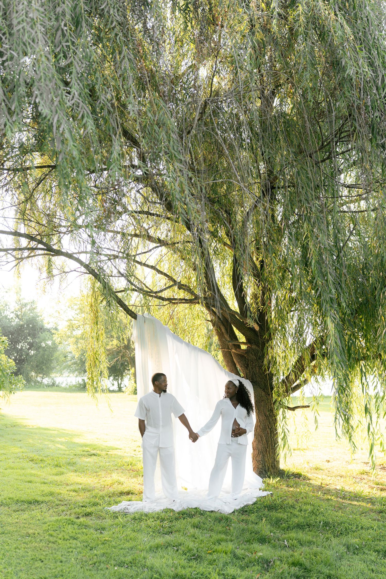 Couple in white outfits holding hands under a weeping willow tree in a sunny green park.