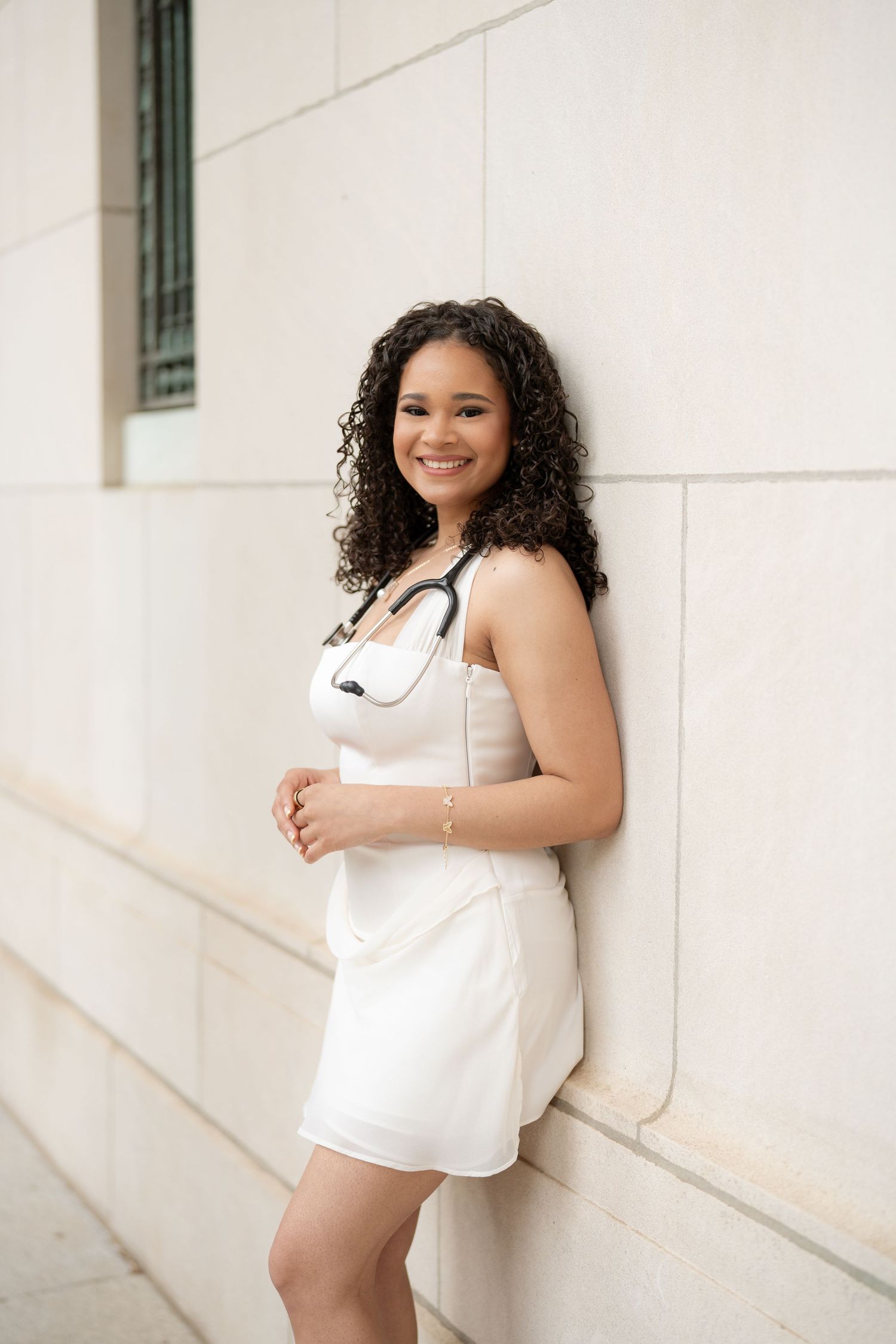 Smiling young woman in white dress with stethoscope leaning against a light stone wall outdoors.