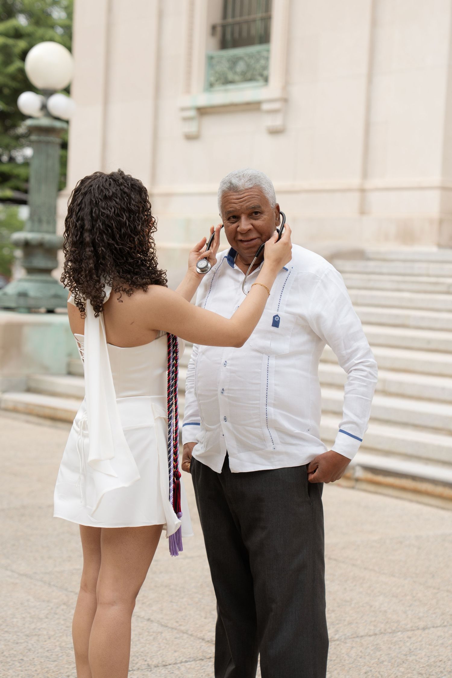 Young woman in white graduation dress placing stethoscope on smiling older man outside a building.