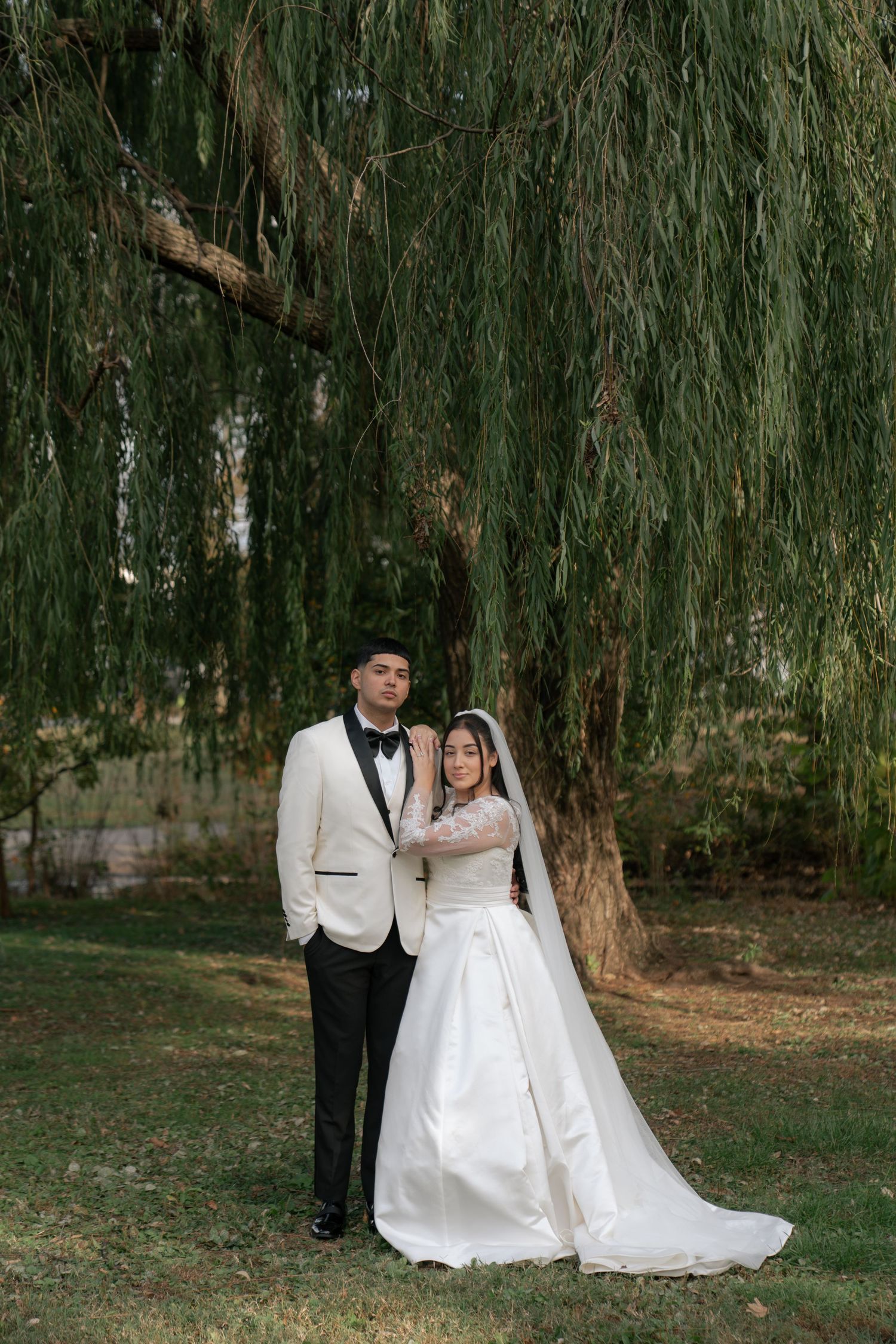 Couple in wedding attire poses under a weeping willow tree on their wedding day.