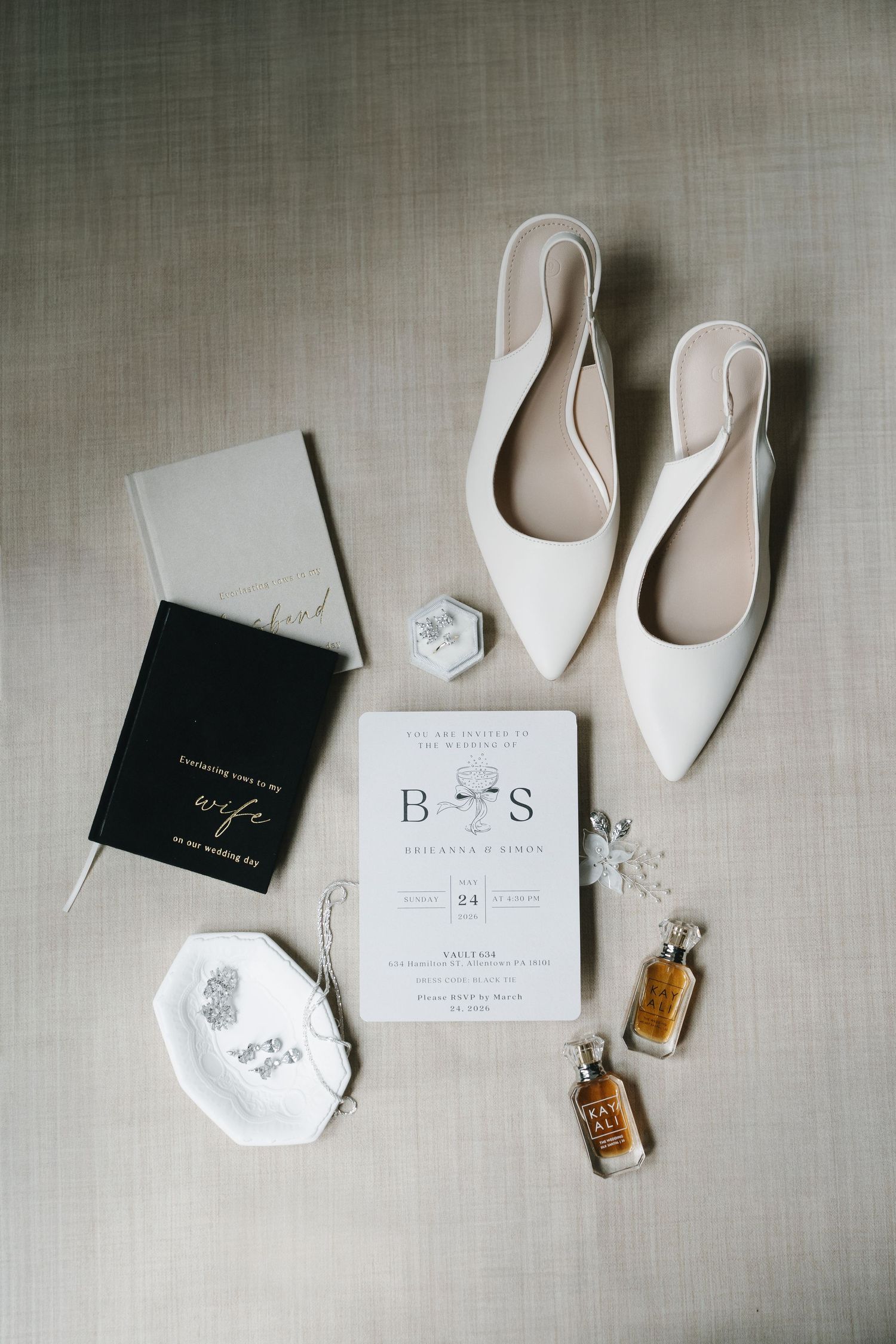 Elegant wedding flat lay with white bridal shoes, monogram invitation, perfume, jewelry, and vow books on neutral background.