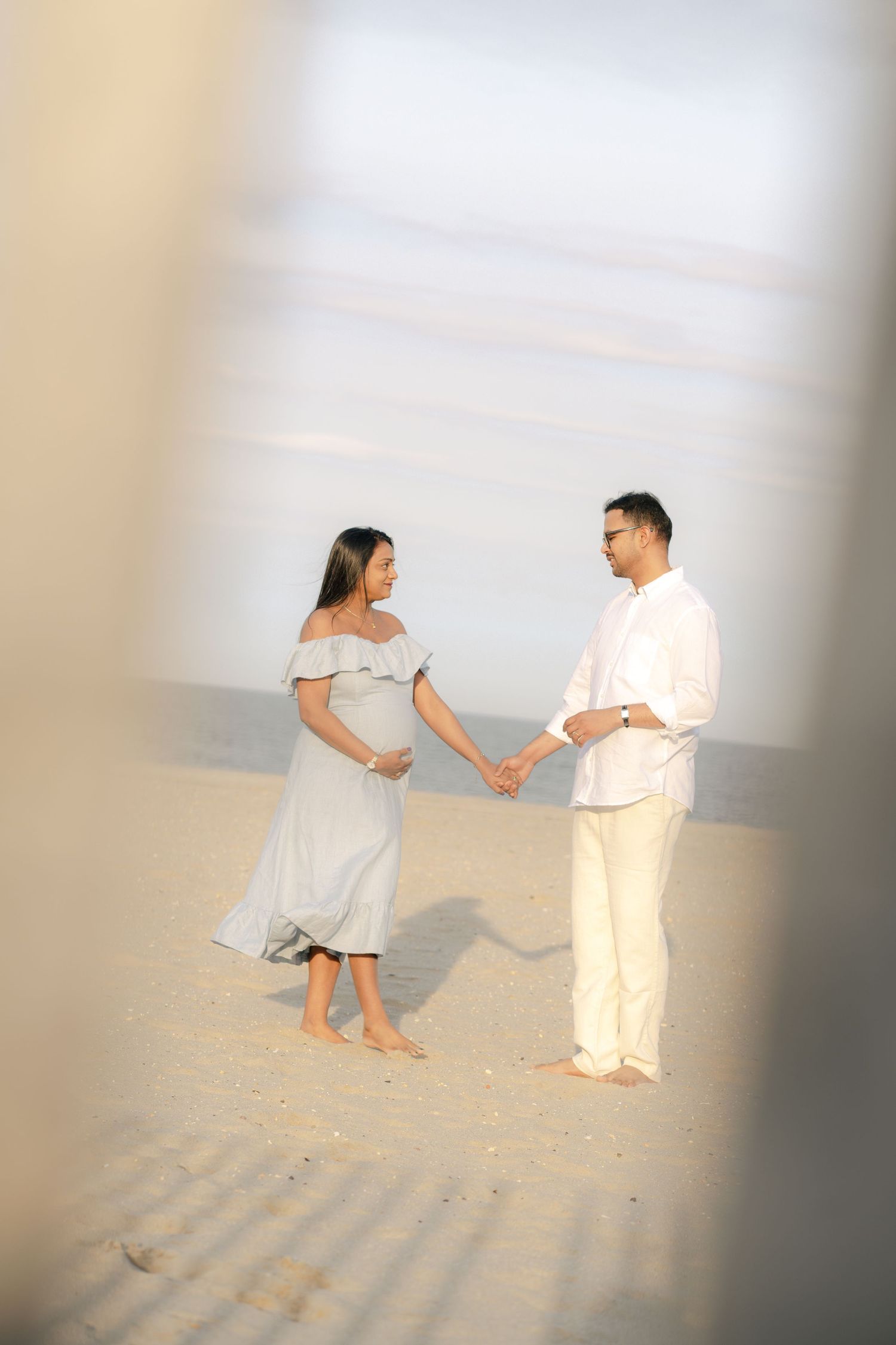 A couple holds hands on a sandy beach, with the person on the left wearing a white off-shoulder dress.