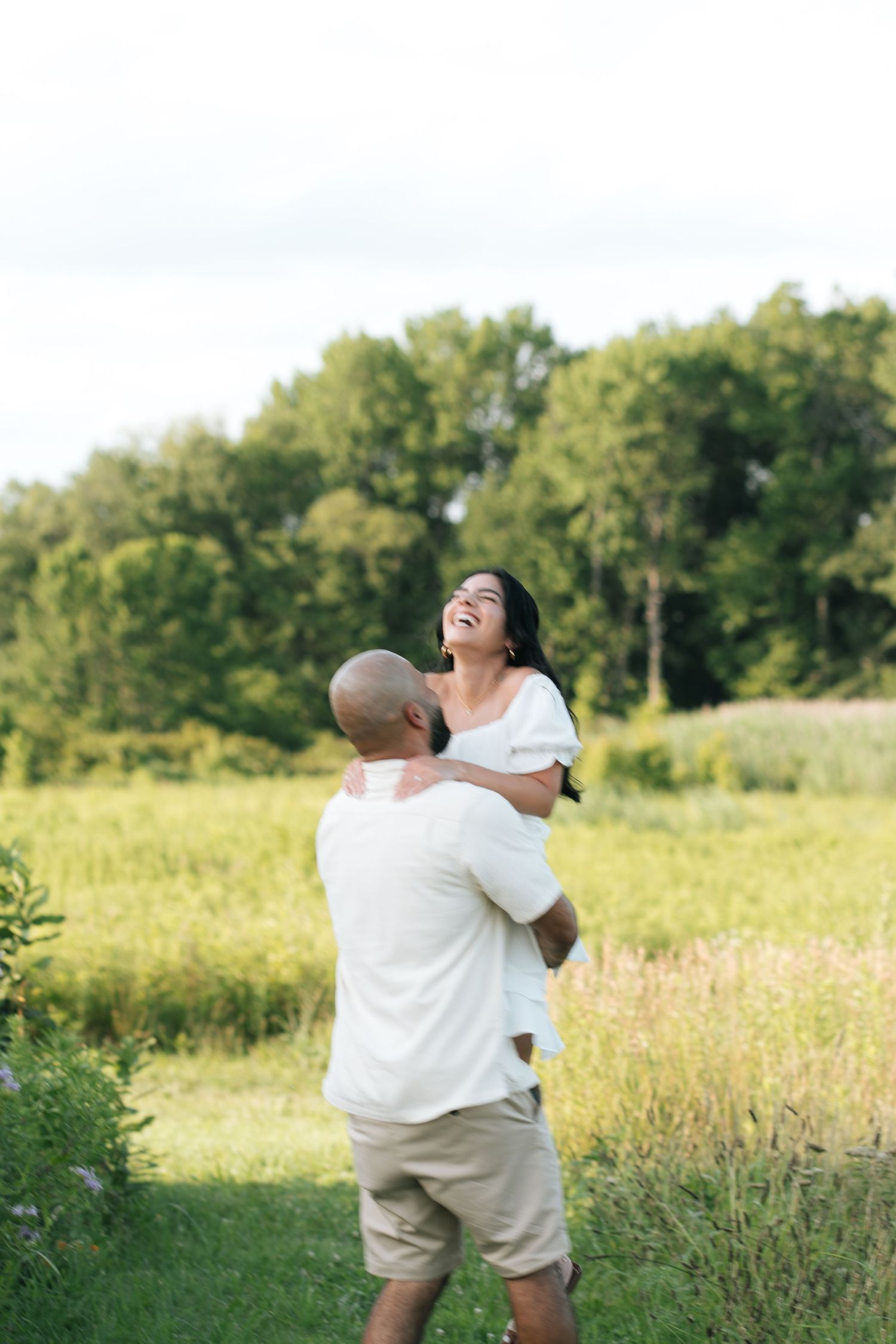A joyful couple embraces in a sunny green meadow, the woman laughing as the man lifts her up outdoors.