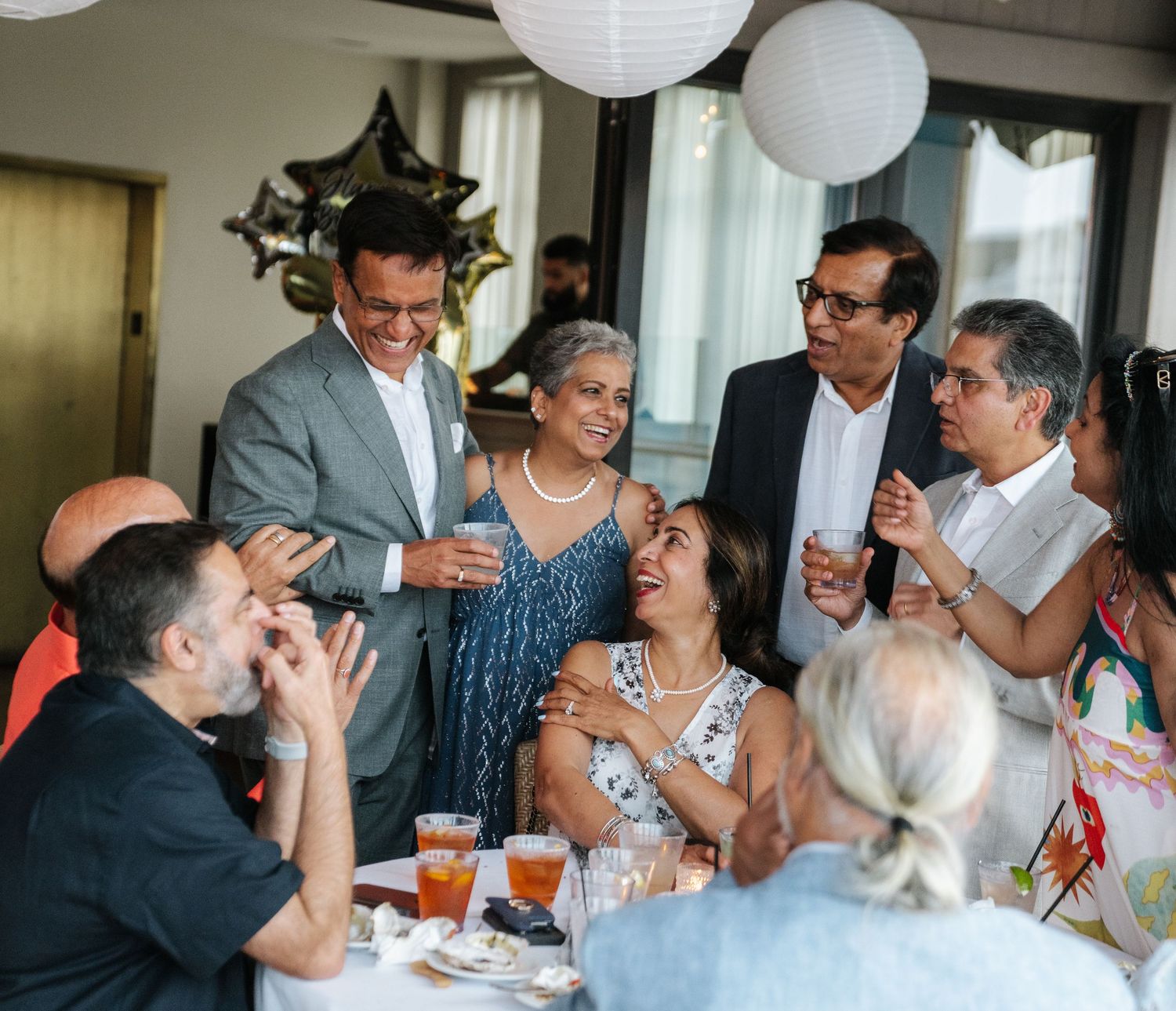 A lively group laughs together at a celebration with white paper lantern decorations overhead.