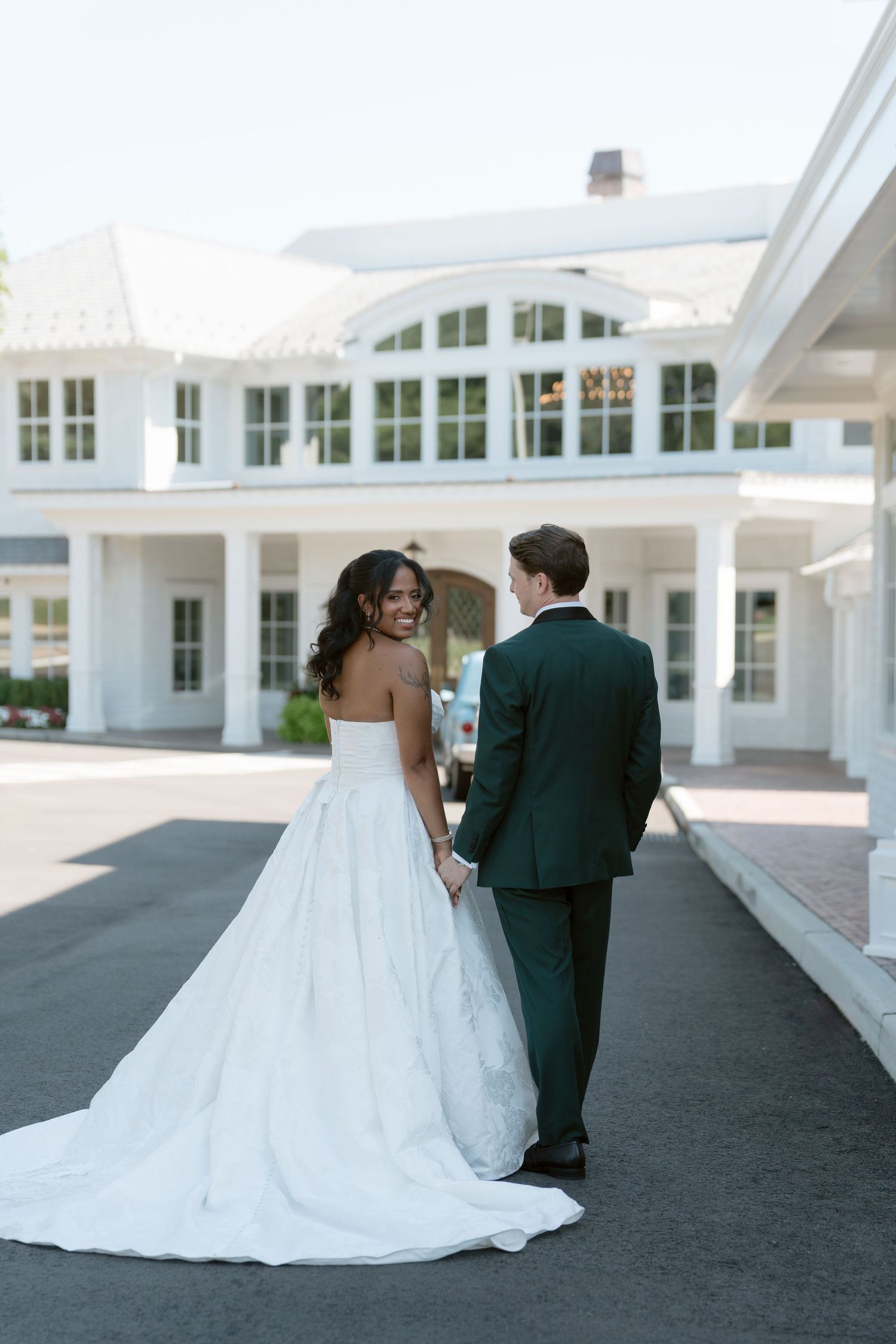 Bride in flowing white gown and groom in dark green suit hold hands outside a white colonial wedding venue.
