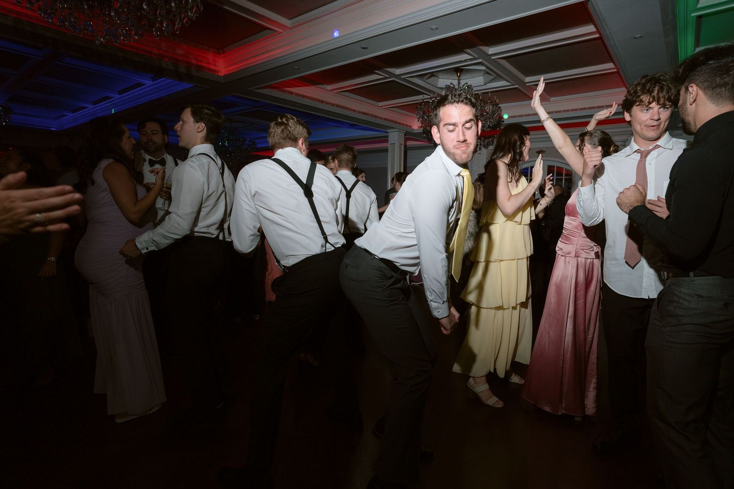 Teens dancing energetically at a formal prom party, wearing suits with suspenders and elegant gowns under colorful lights.